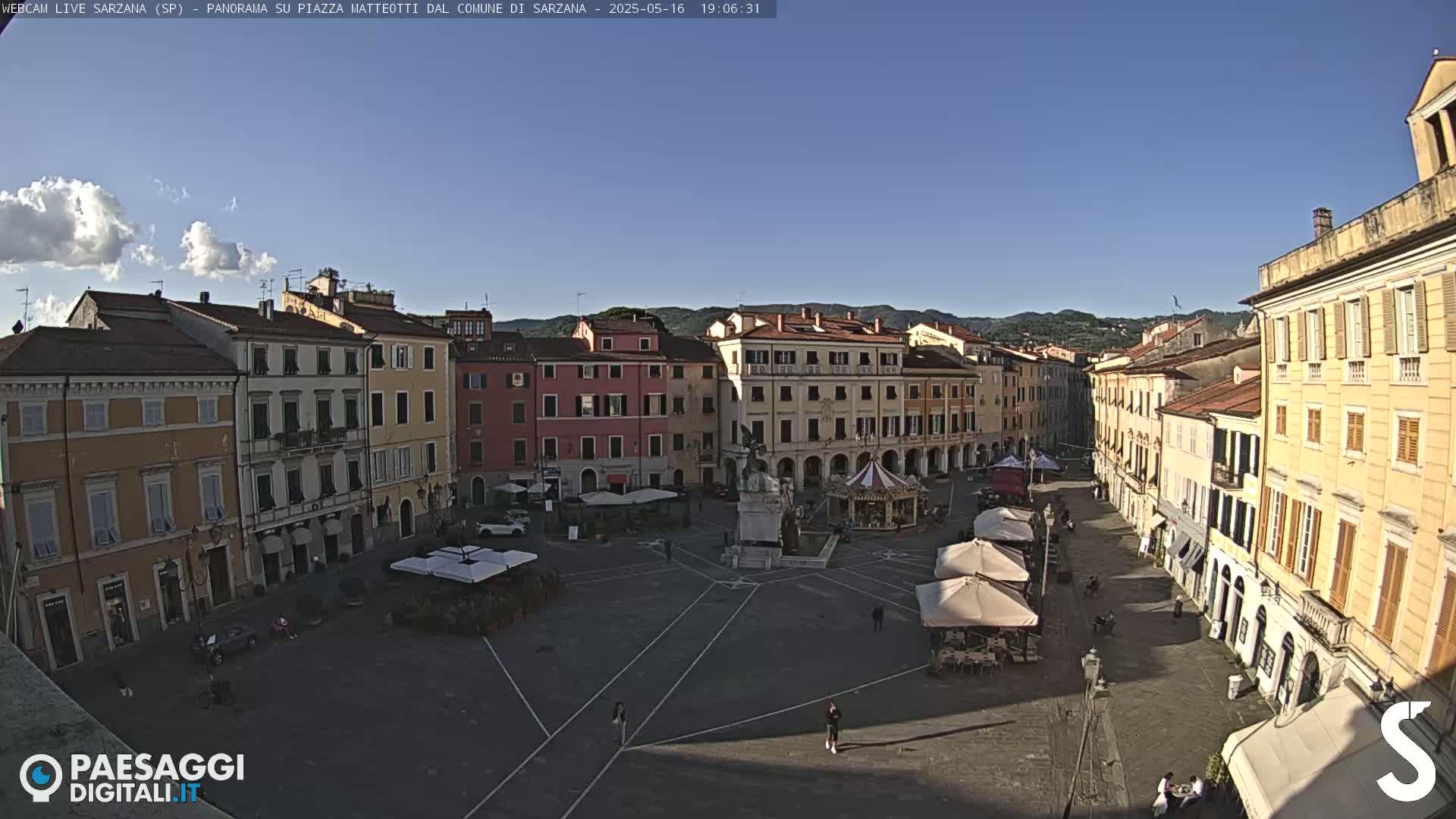 A large, mostly empty town square with surrounding buildings under a clear, sunny sky.