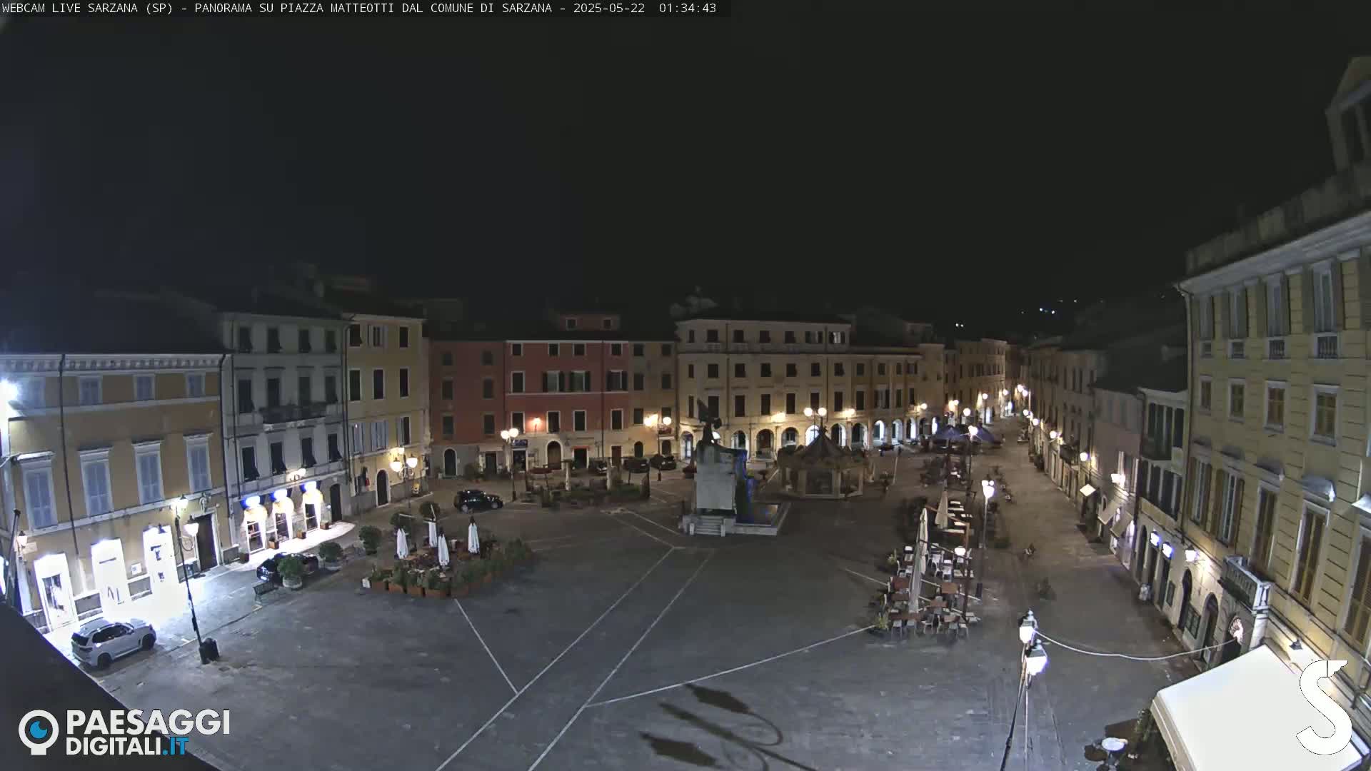 A nighttime, wide shot of a town square with several buildings surrounding it, a few cars parked, and some outdoor seating areas.