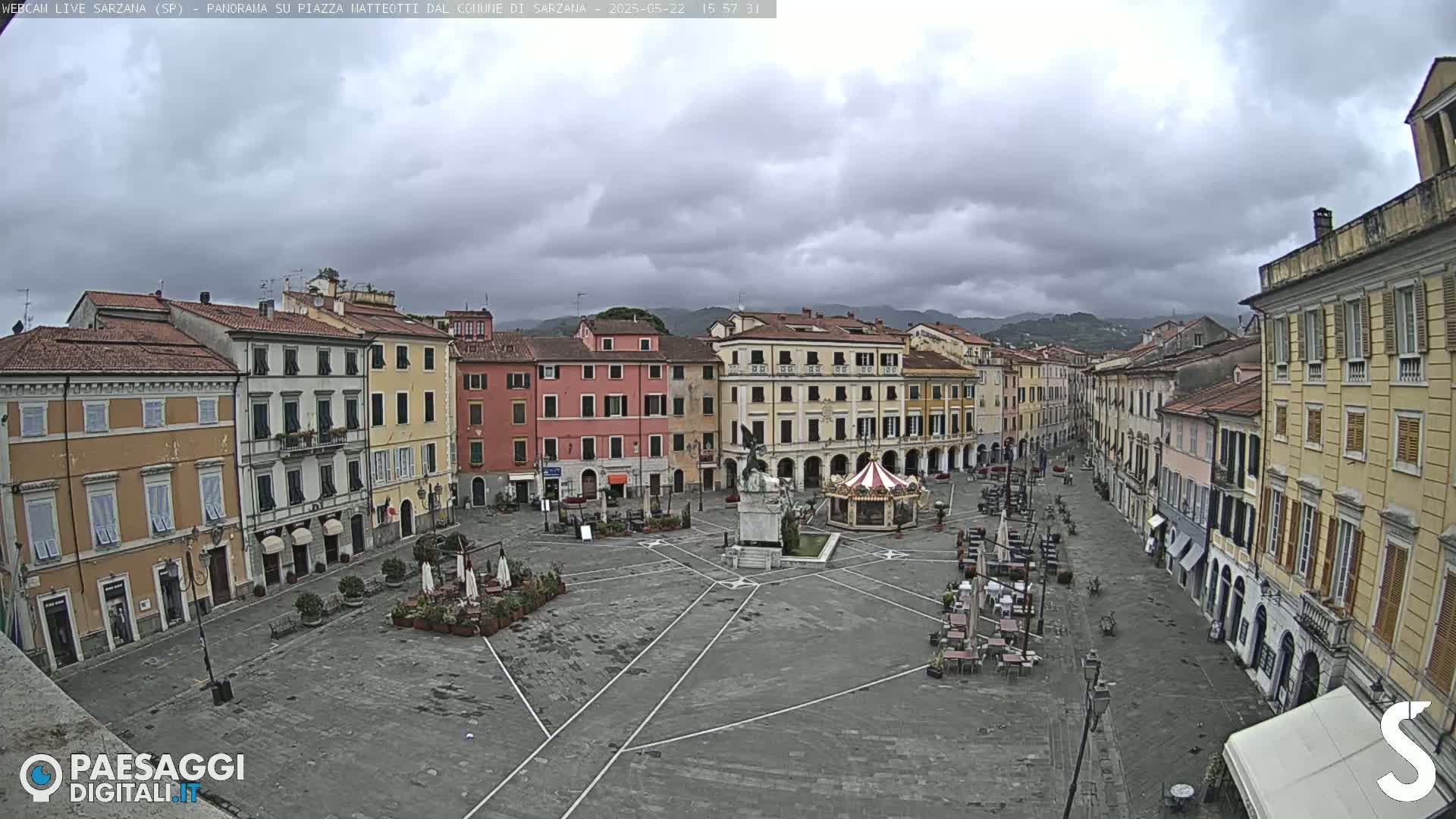A mostly empty town square, under a cloudy sky, is surrounded by multi-story buildings.