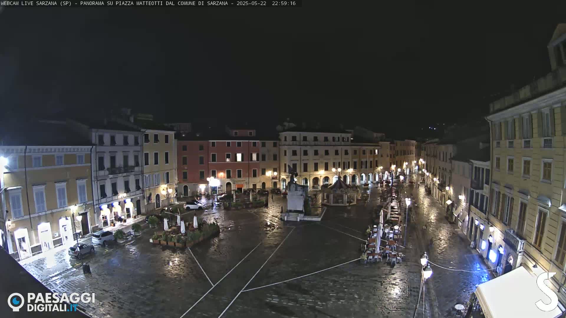 A wet, nighttime view of a town square, surrounded by buildings with illuminated windows and outdoor seating areas.