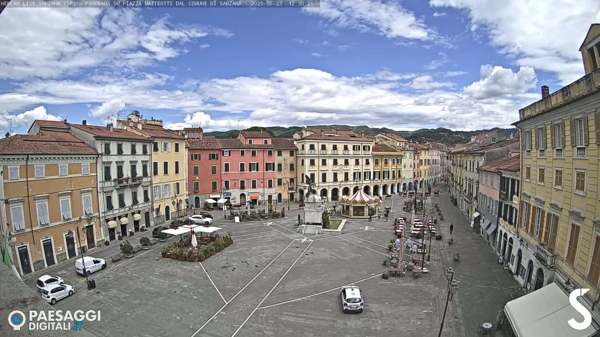 A partly cloudy day reveals a large town square surrounded by multi-story buildings, with a few cars and outdoor seating areas visible.
