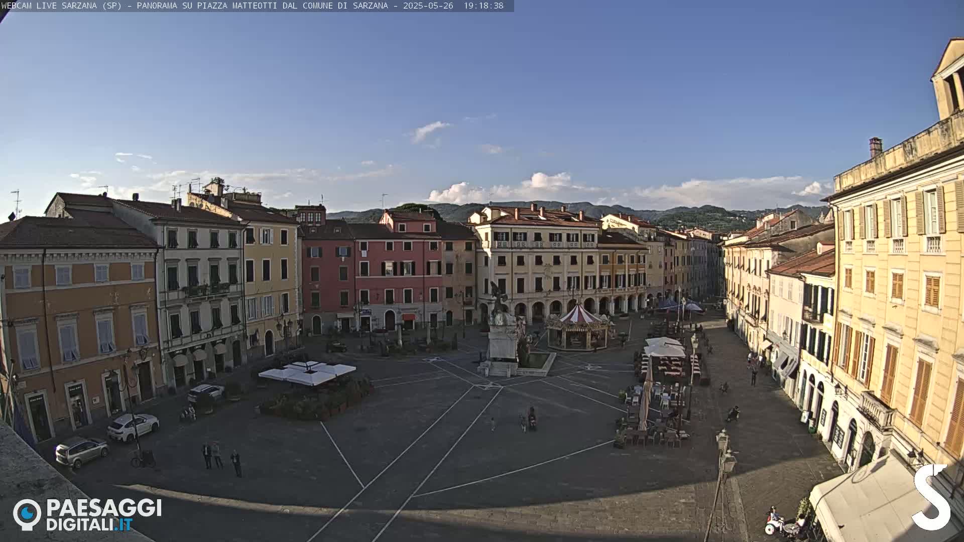 A mostly sunny town square with several multi-story buildings surrounding it, a carousel, and a few people walking around.