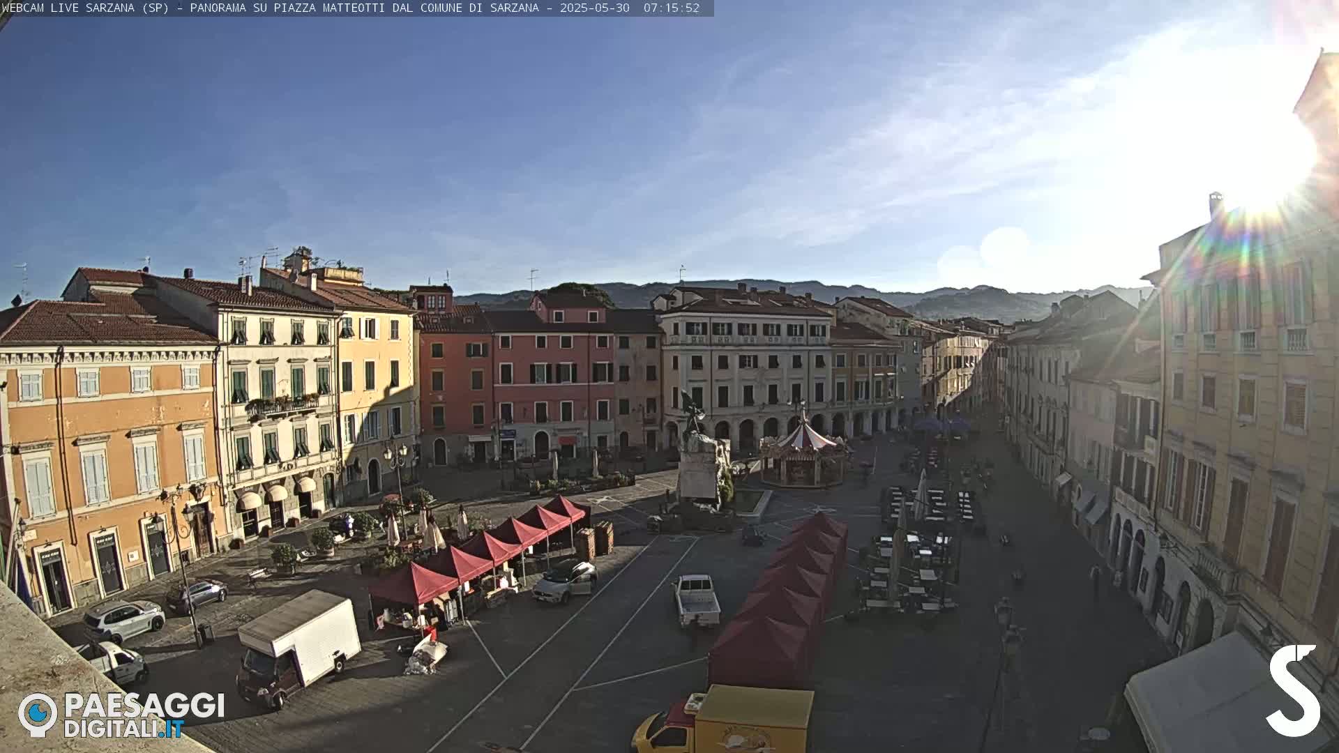 A sunny morning view of a town square surrounded by pastel-colored buildings, with a few cars and market stalls present.