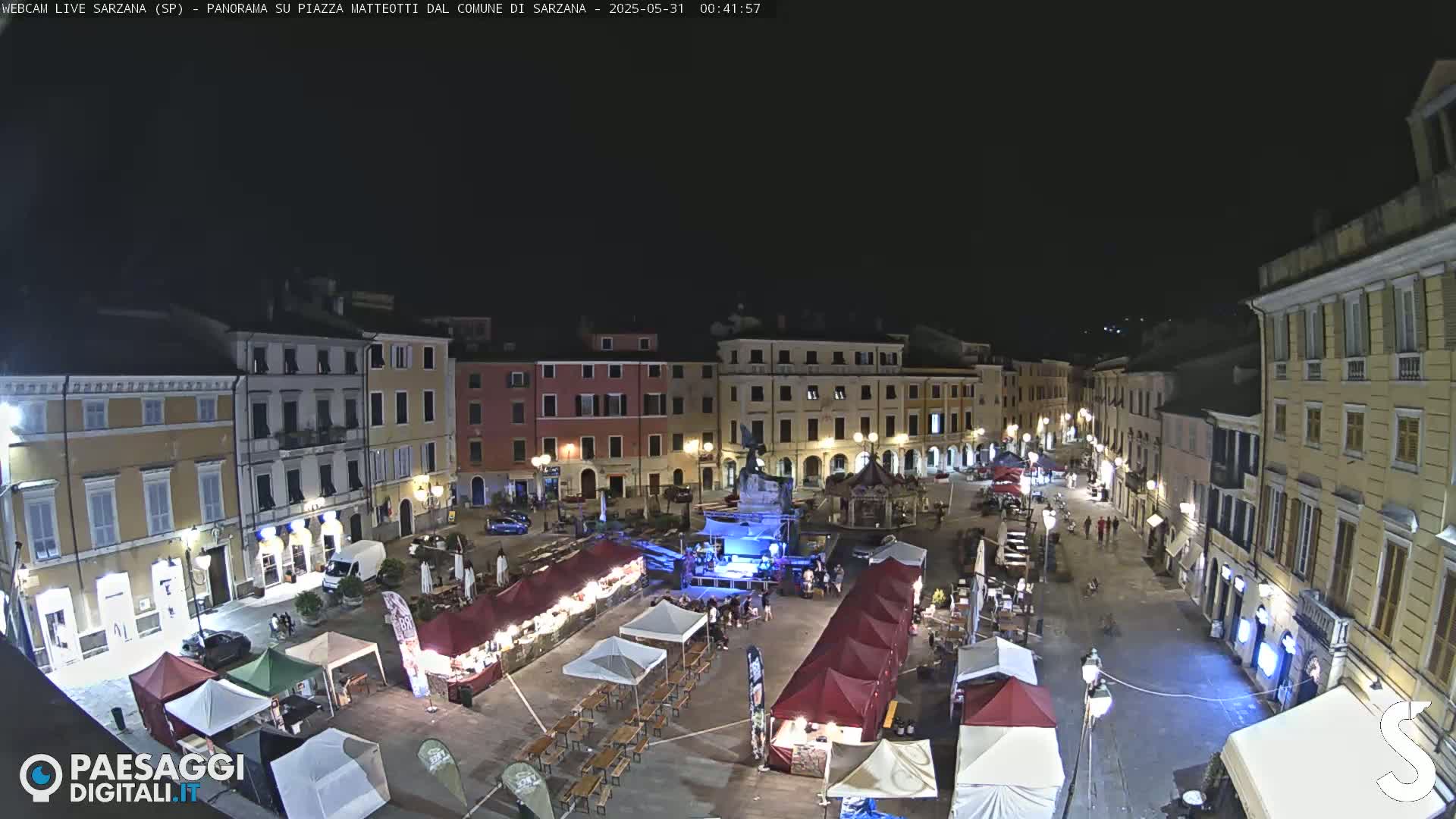A nighttime view of a town square filled with market stalls and people, under a clear, dark sky.