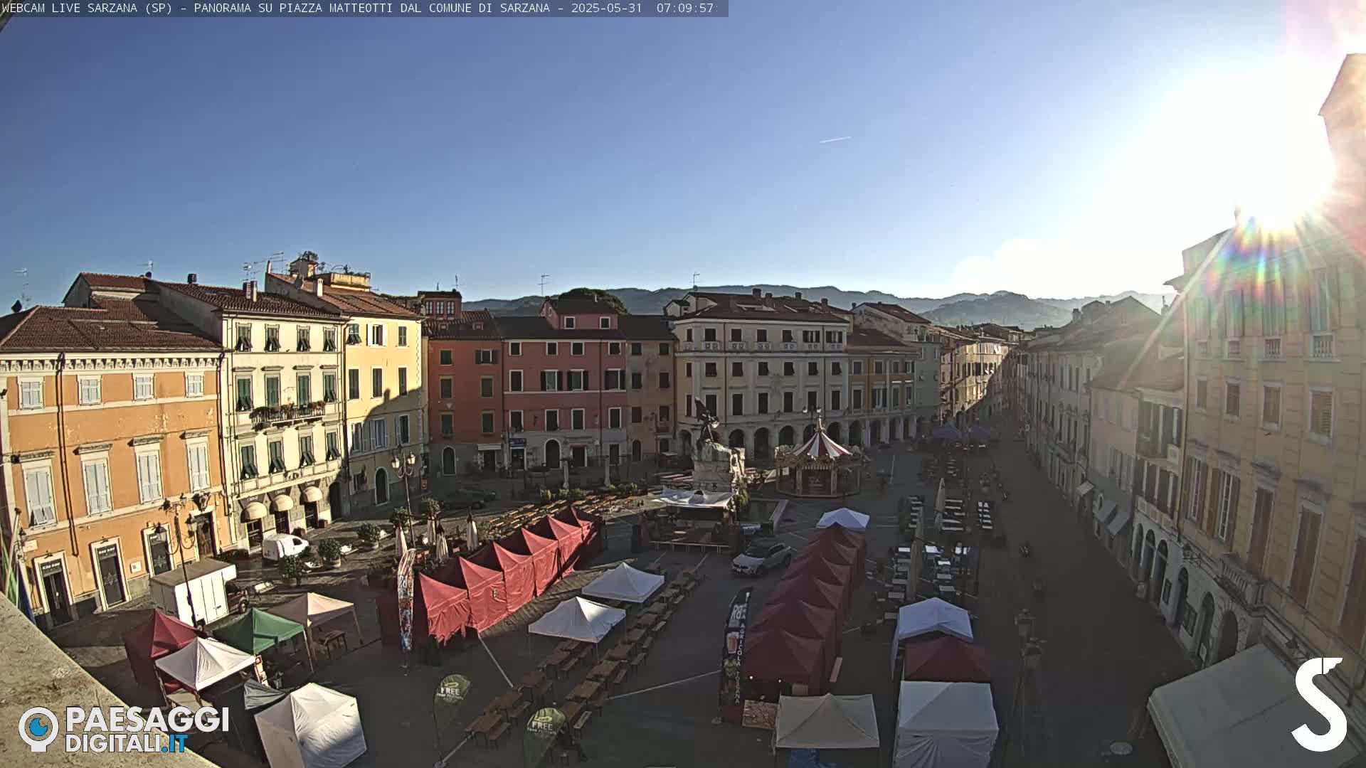 A sunny day reveals a town square filled with market stalls and surrounded by historical buildings, with mountains visible in the background.