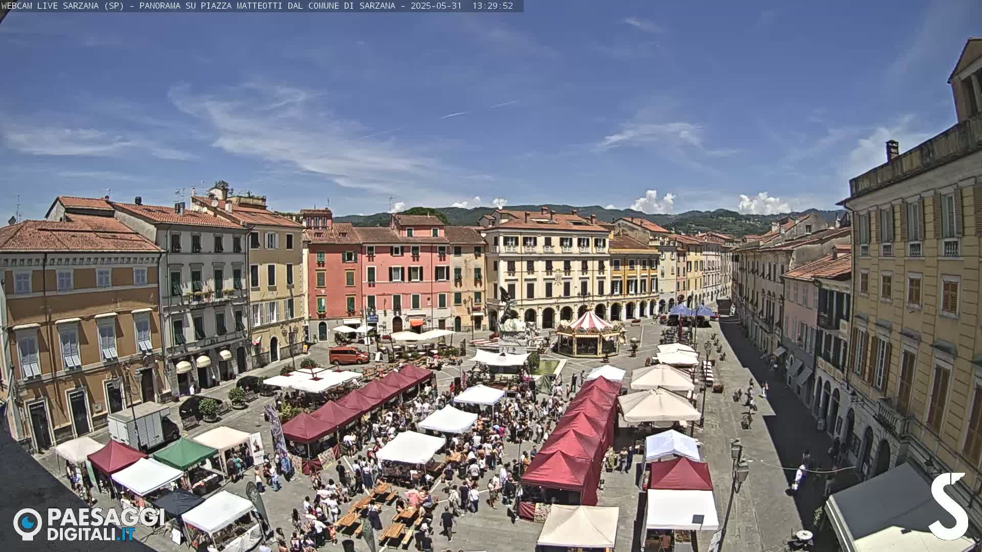 A sunny day shows a large town square filled with market stalls and people, surrounded by historic buildings.