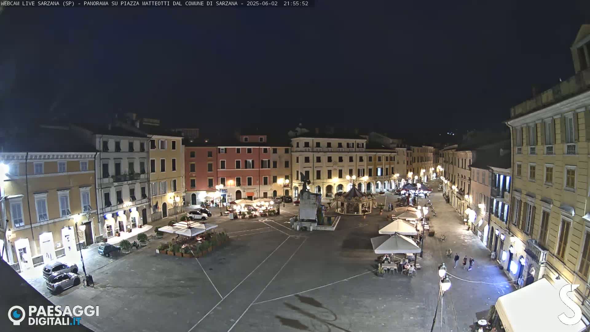 A nighttime view of a town square, illuminated by streetlights, with several buildings surrounding it and a few people walking around.