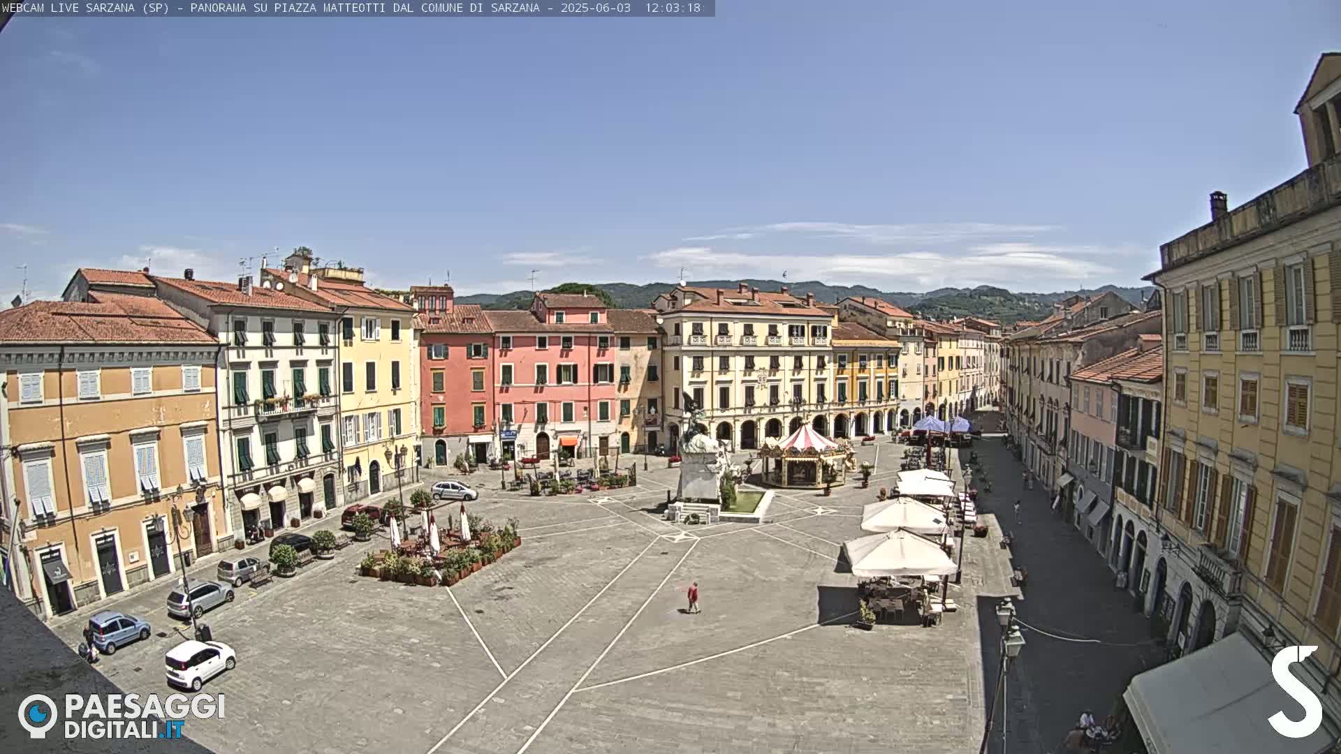 A mostly sunny day reveals a large town square surrounded by multi-story buildings, a central monument, and a few parked cars.
