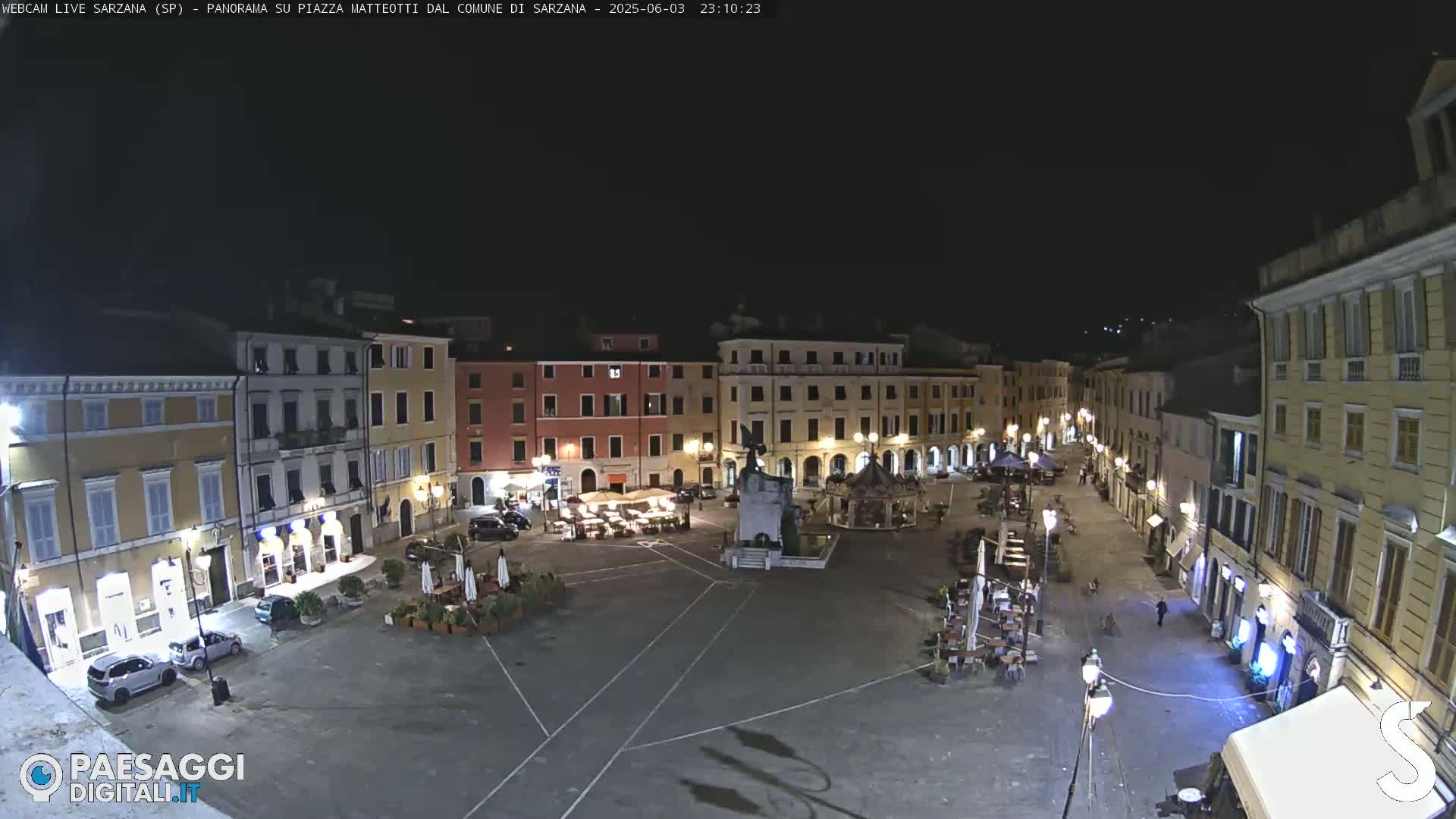 A nighttime view of a town square, illuminated by streetlights, with several buildings surrounding it and a few people and cars visible.