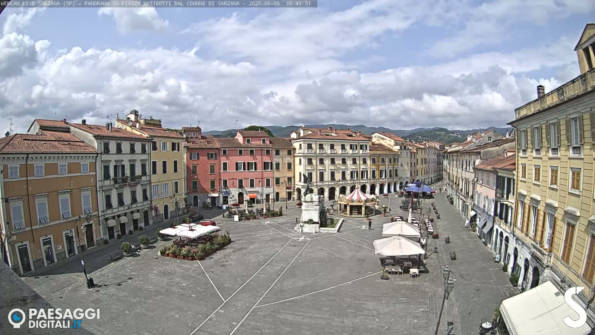 A partly cloudy day reveals a large town square surrounded by multi-story buildings, with a few outdoor cafes and a carousel.