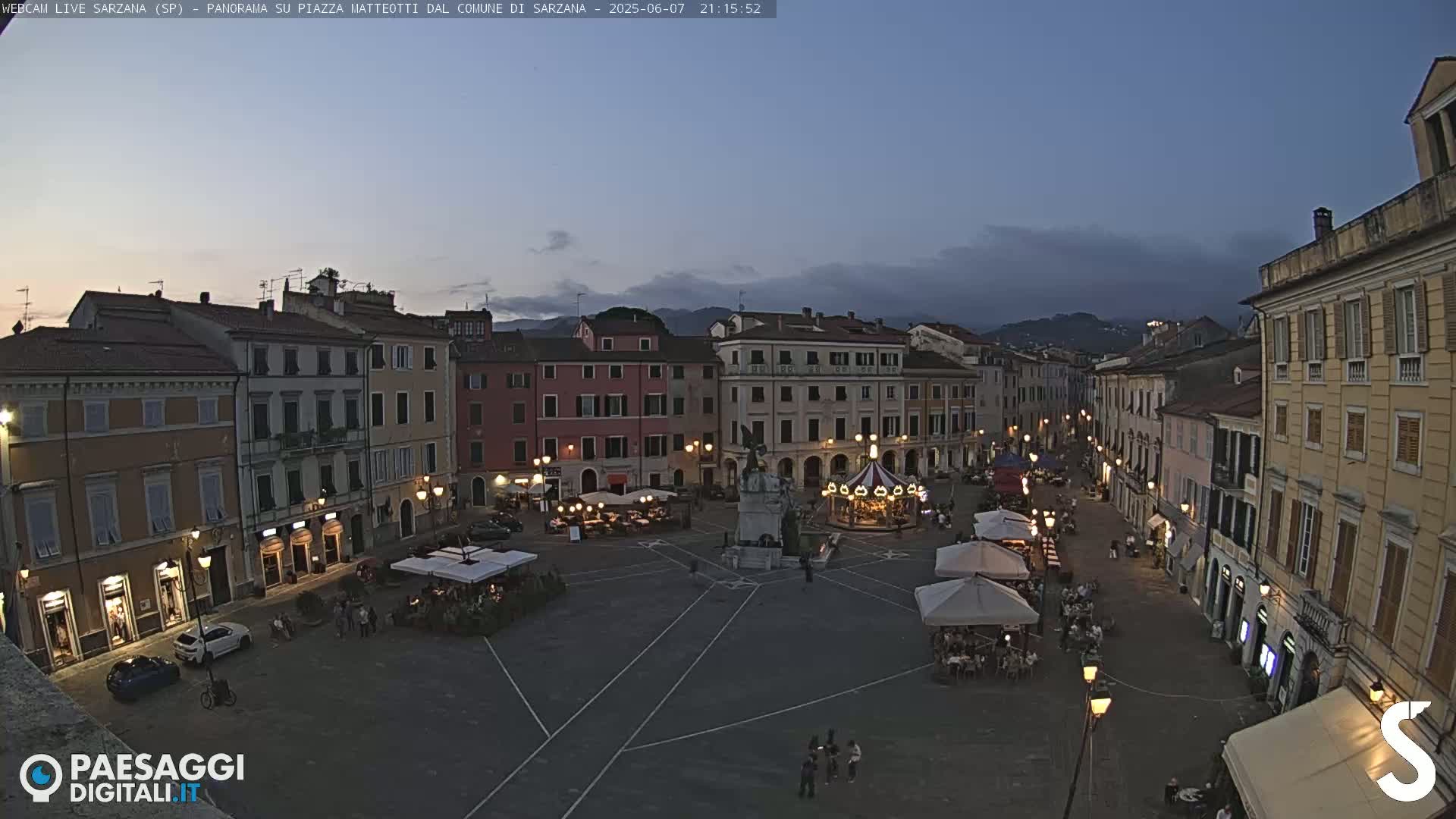 A twilight view of a town square with several buildings surrounding it, a carousel, and outdoor seating areas under umbrellas.