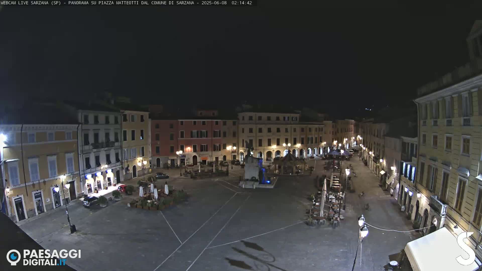 A nighttime, wide shot of a town square with several buildings surrounding it, a fountain in the center, and outdoor seating areas.