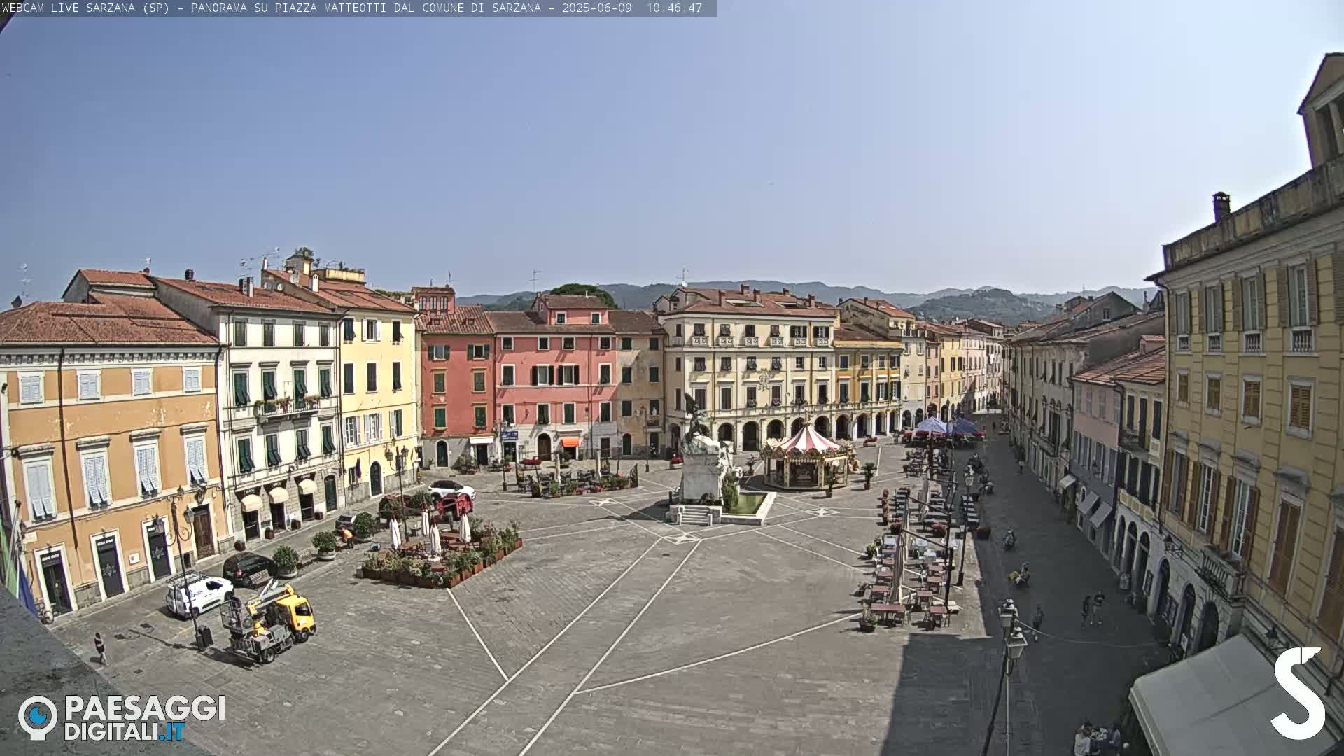 A large, paved town square is surrounded by multi-story buildings under a clear, sunny sky.