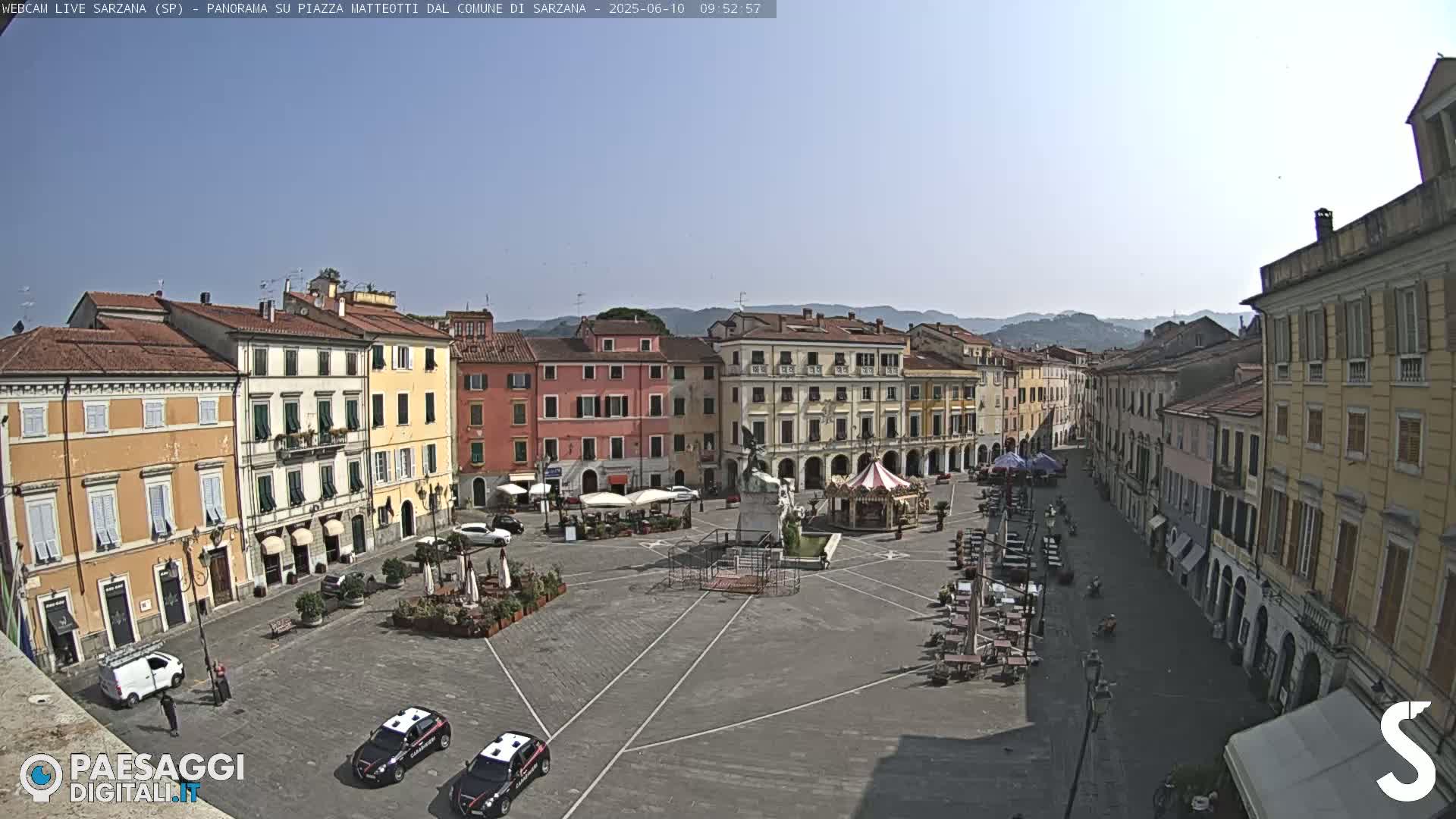 A sunny day in a large town square surrounded by multi-story buildings, with a few cars and outdoor seating visible.