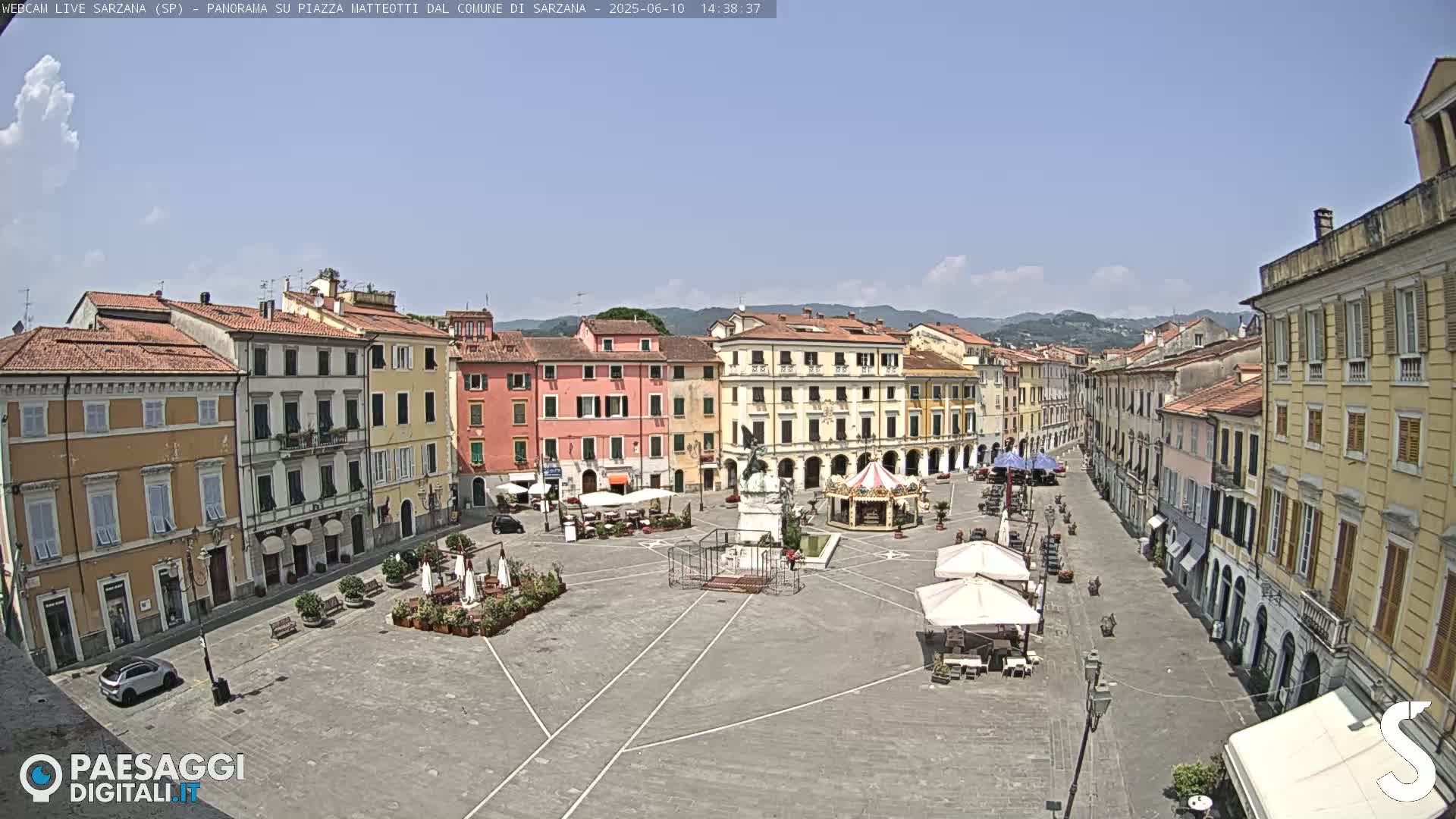 A mostly sunny day reveals a large town square, surrounded by multi-story buildings, with a few people and some outdoor seating under umbrellas.
