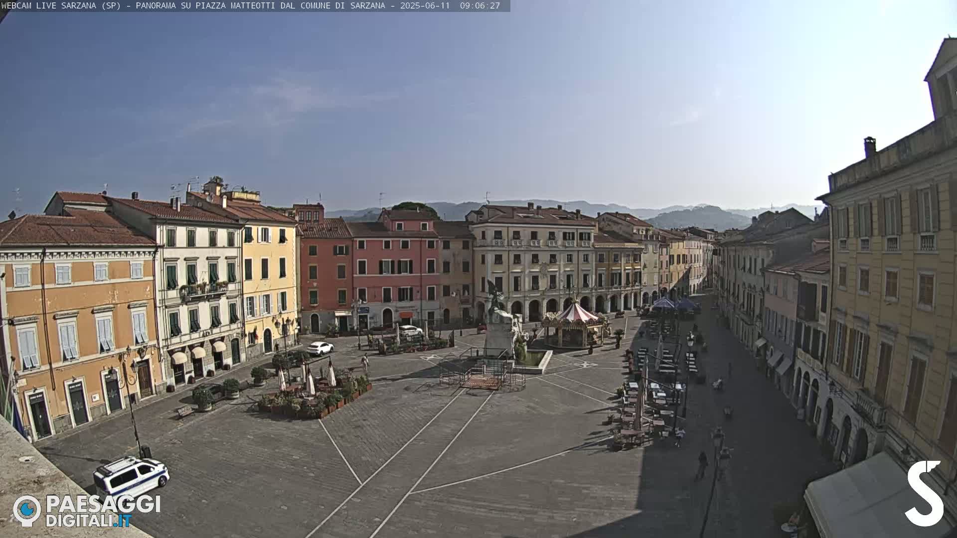 A mostly empty town square, surrounded by multi-story buildings, under a clear, sunny sky.