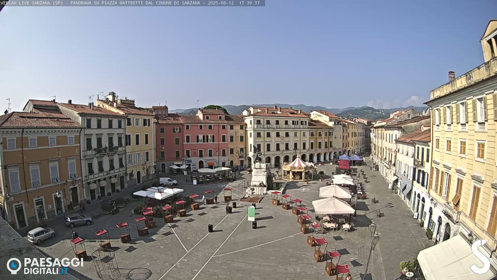 A town square in Italy, under a clear blue sky, features several multi-story buildings surrounding the square which is partially occupied by outdoor seating and a small carousel.