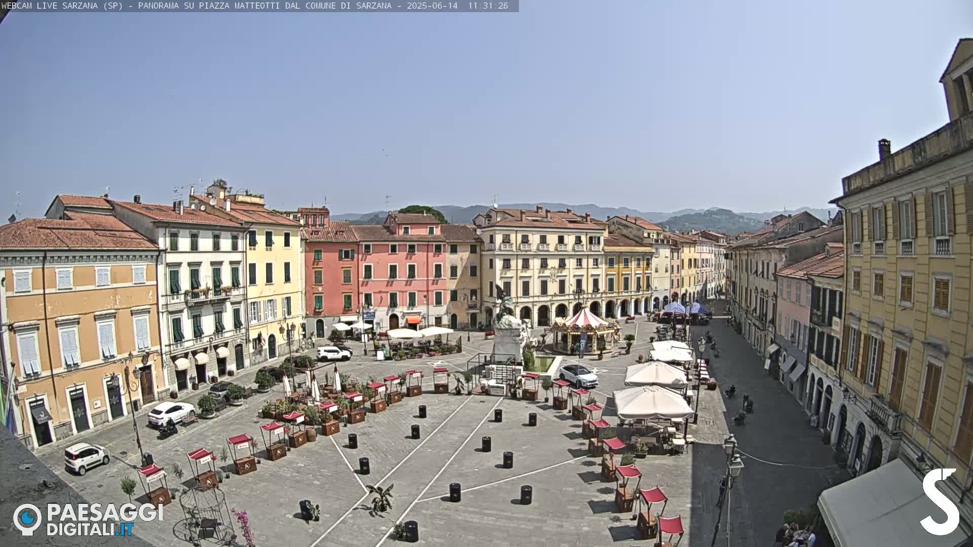 A town square in bright sunlight, surrounded by multi-story buildings, features several market stalls and outdoor seating areas.