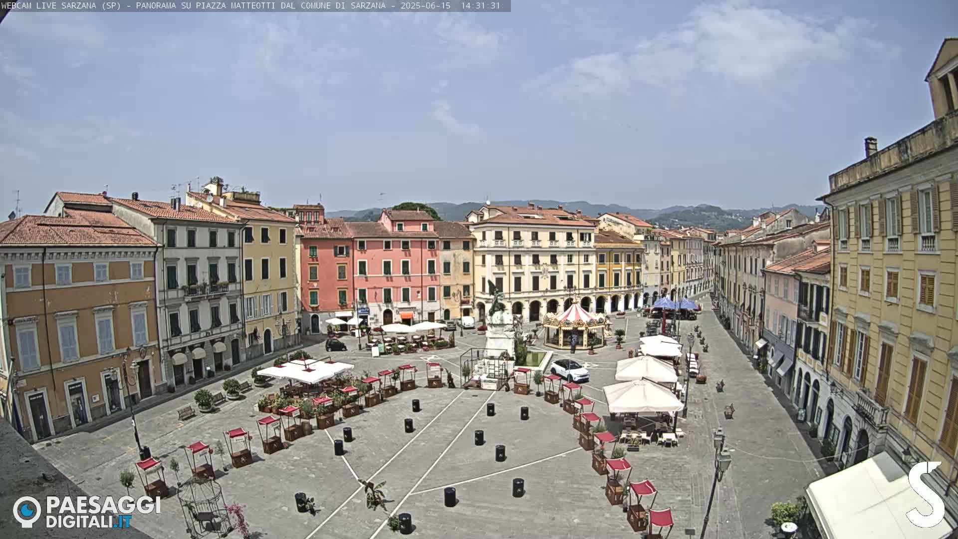 A mostly sunny day reveals a large town square surrounded by multi-story buildings, featuring several outdoor market stalls and a carousel.