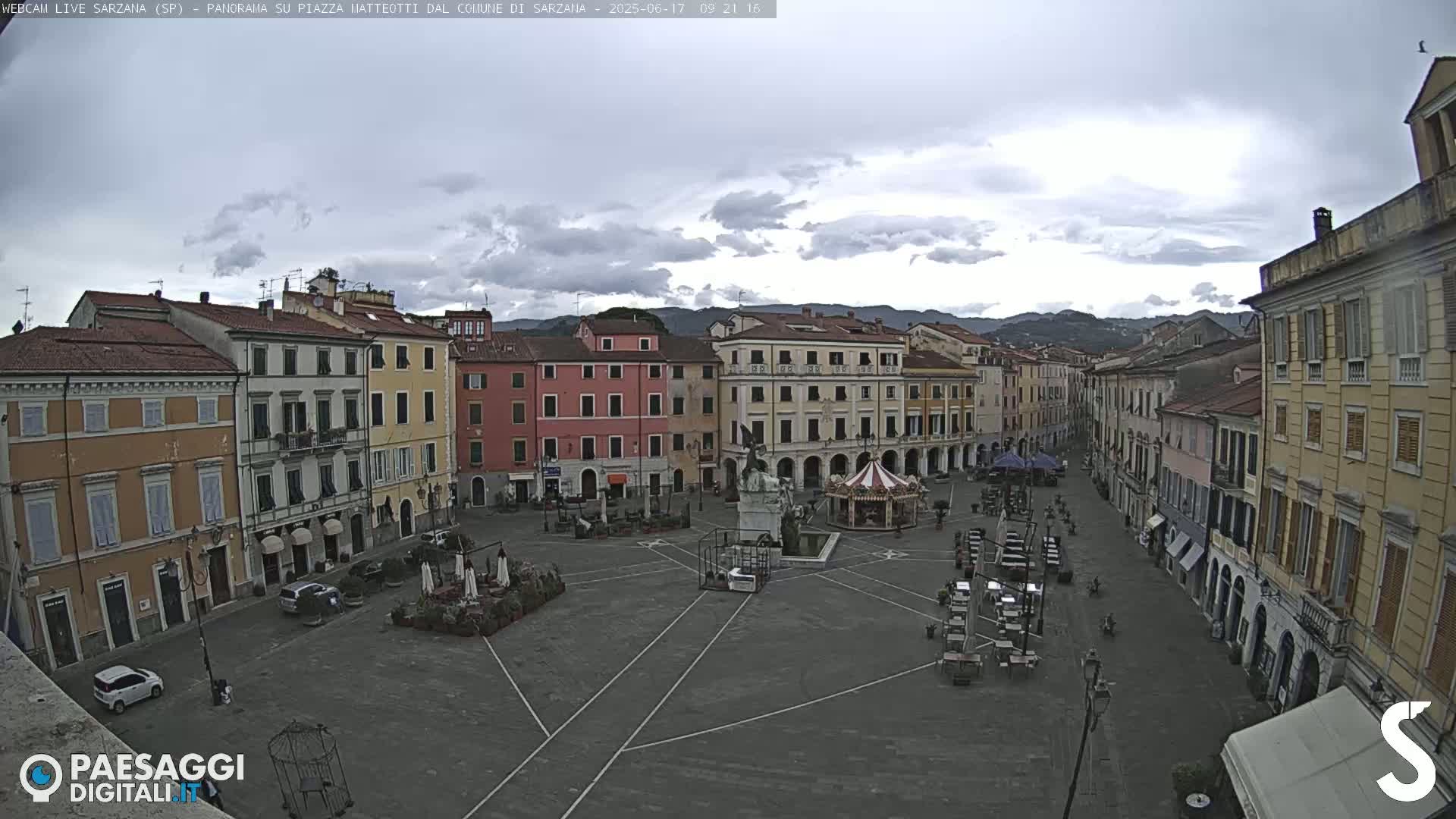 A mostly empty town square with multi-story buildings surrounding it under an overcast sky.
