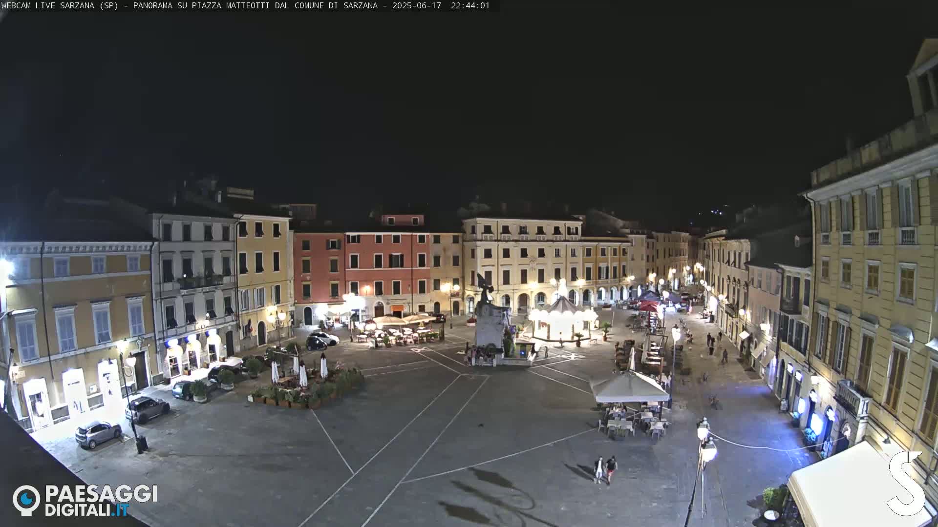 A nighttime view of a town square with several buildings surrounding it, some with outdoor seating areas, under a clear, dark sky.