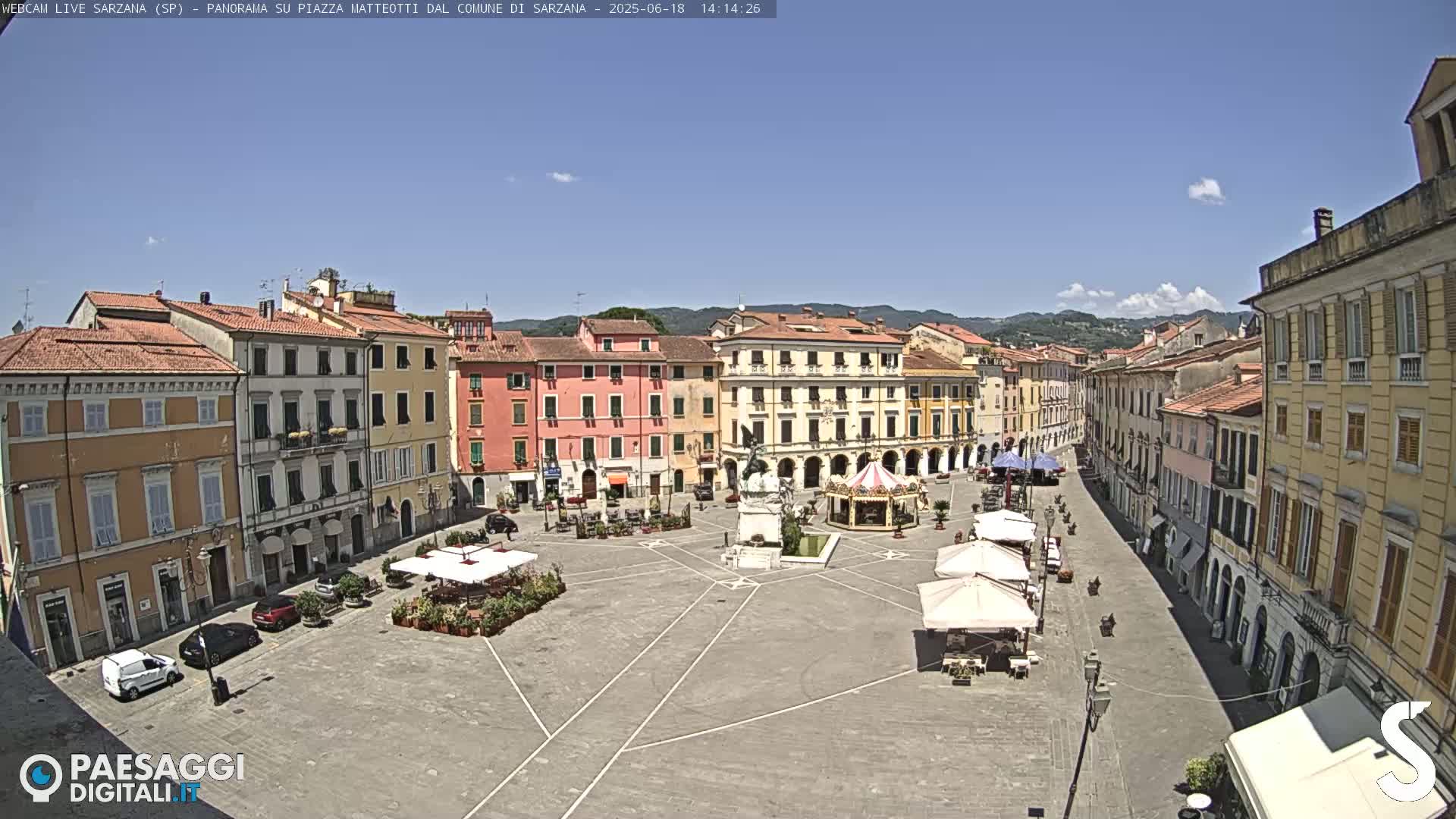 A sunny day reveals a large town square in Italy, surrounded by multi-story buildings with arched walkways, a few cars parked, and outdoor seating under umbrellas.