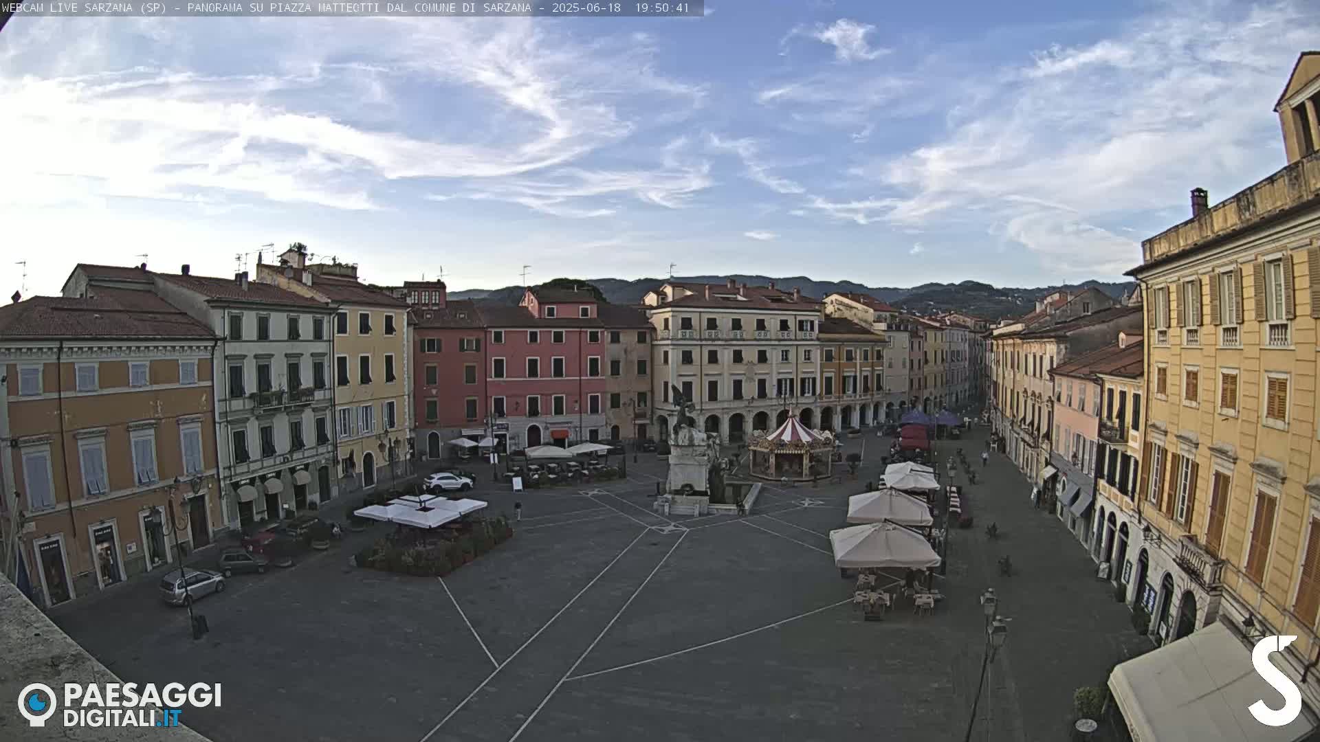 A mostly sunny, partly cloudy day overlooks a large town square in Italy, where several multi-story buildings surround a paved plaza with a few cars and outdoor seating areas under umbrellas.