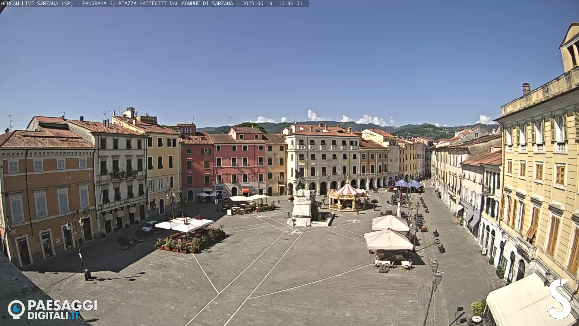 A sunny, mostly clear day reveals a large town square surrounded by multi-story buildings, featuring a monument and carousel in the center, and several outdoor cafe umbrellas.