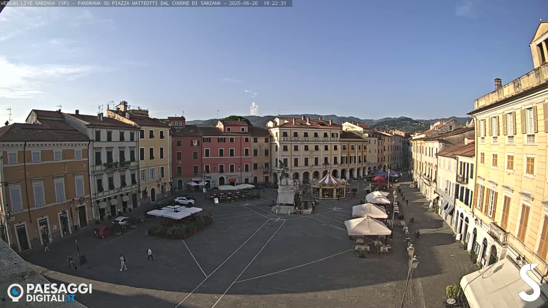 A mostly sunny town square with multi-story buildings surrounding it, a monument and carousel in the center, and outdoor seating at cafes.