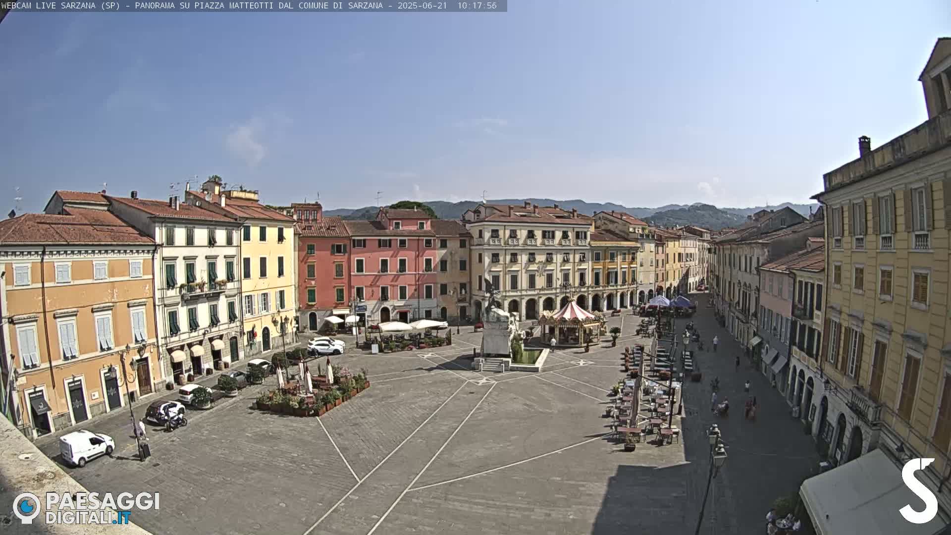 A large, paved town square is surrounded by multi-story buildings under a clear, sunny sky.