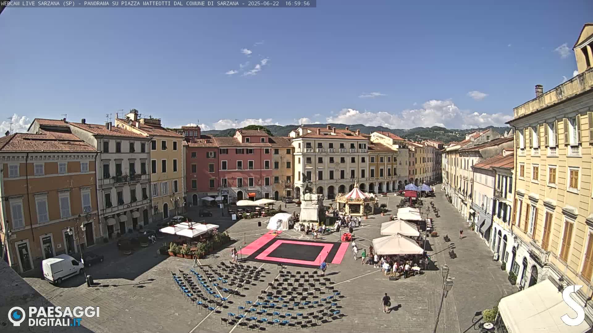 A sunny day shows a town square surrounded by buildings, with a black-covered area, several white umbrellas, and rows of empty chairs set up in the plaza.