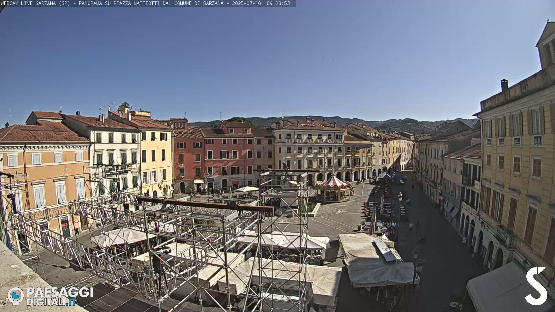 A large town square, under a clear blue sky, is filled with temporary market stalls and scaffolding, surrounded by multi-story buildings.