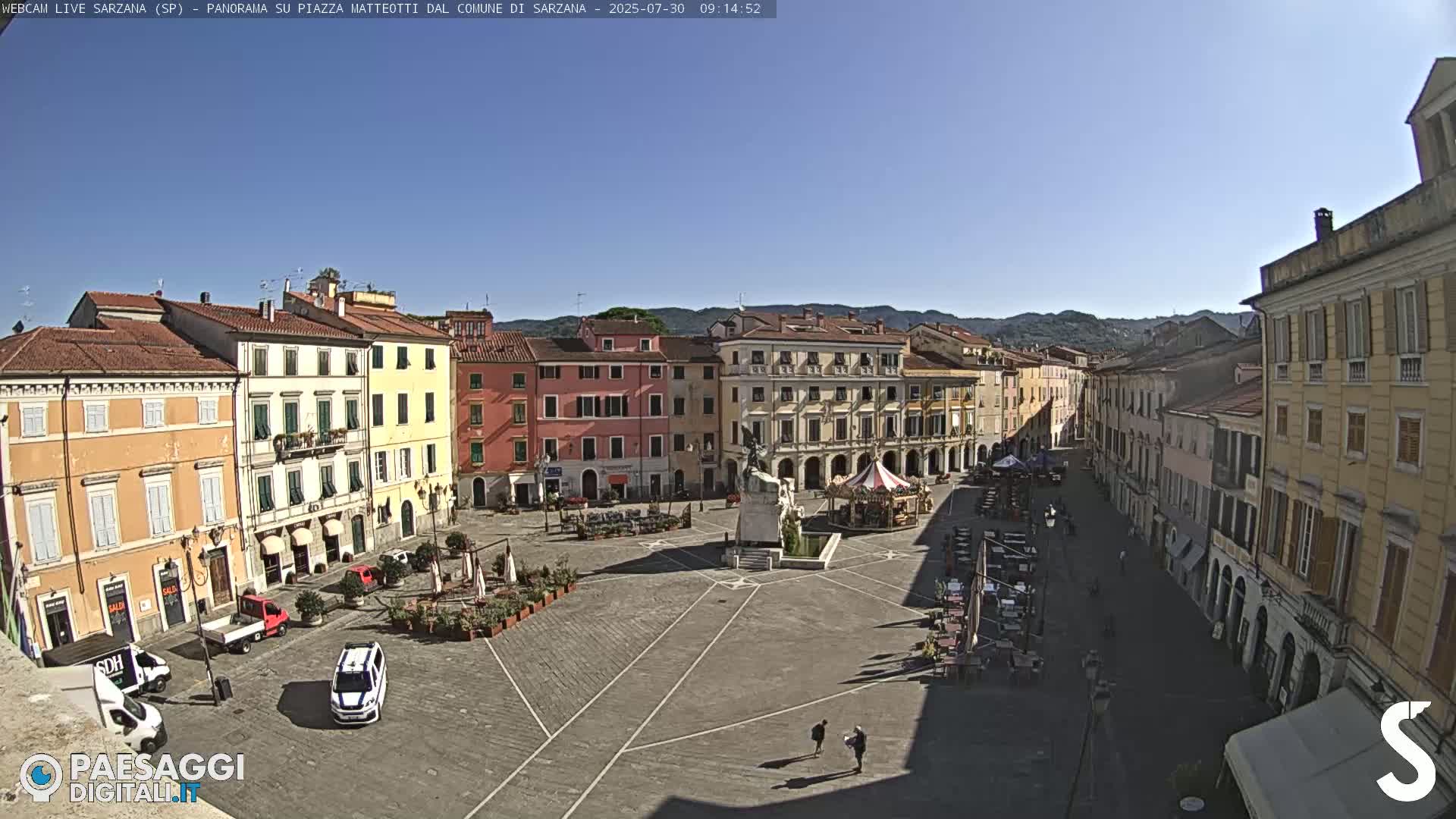A large, paved town square surrounded by multi-story buildings under a clear, sunny sky.