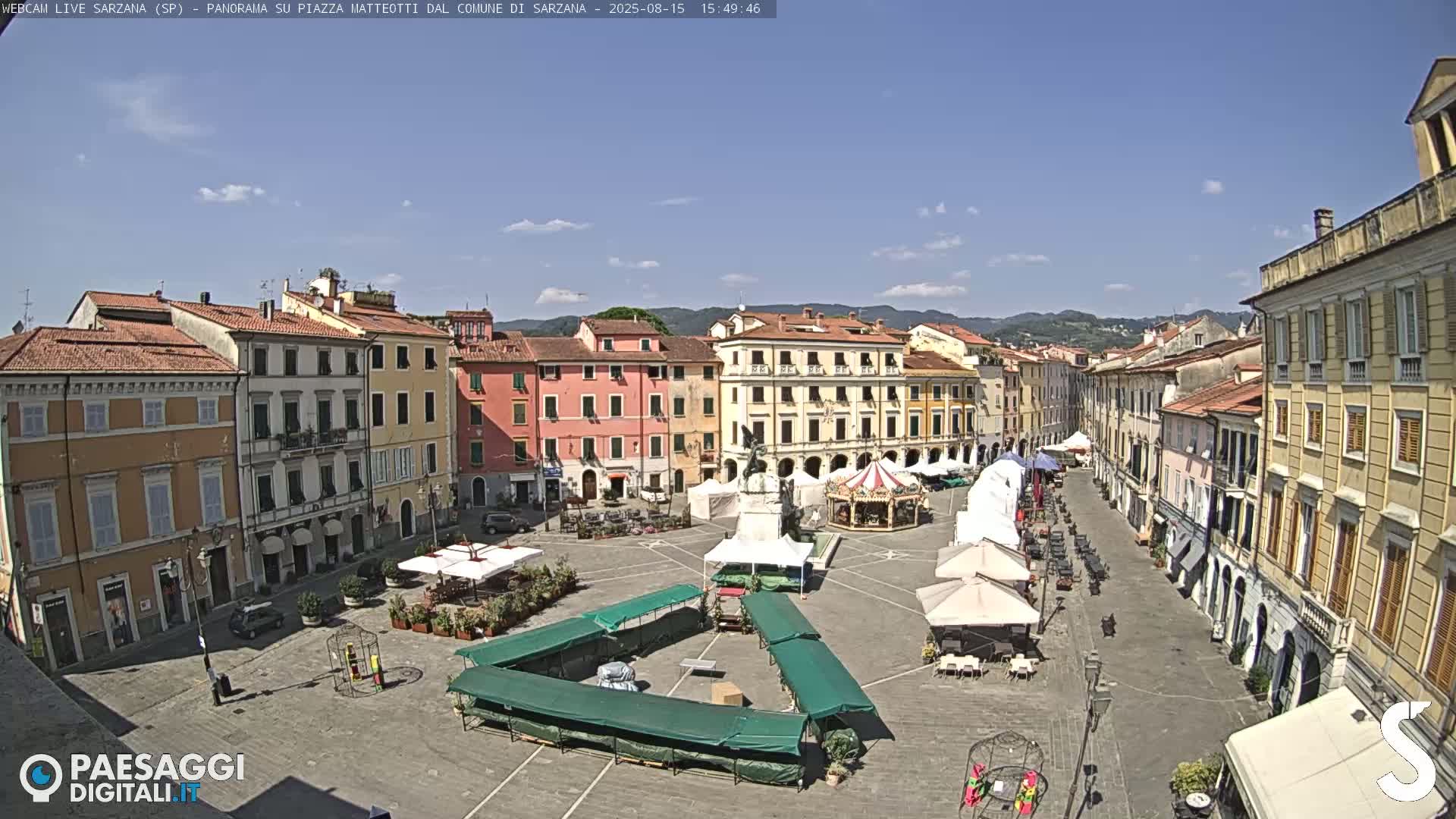 A sunny day reveals a large town square with multiple buildings surrounding it, market stalls set up in the center, and a carousel.