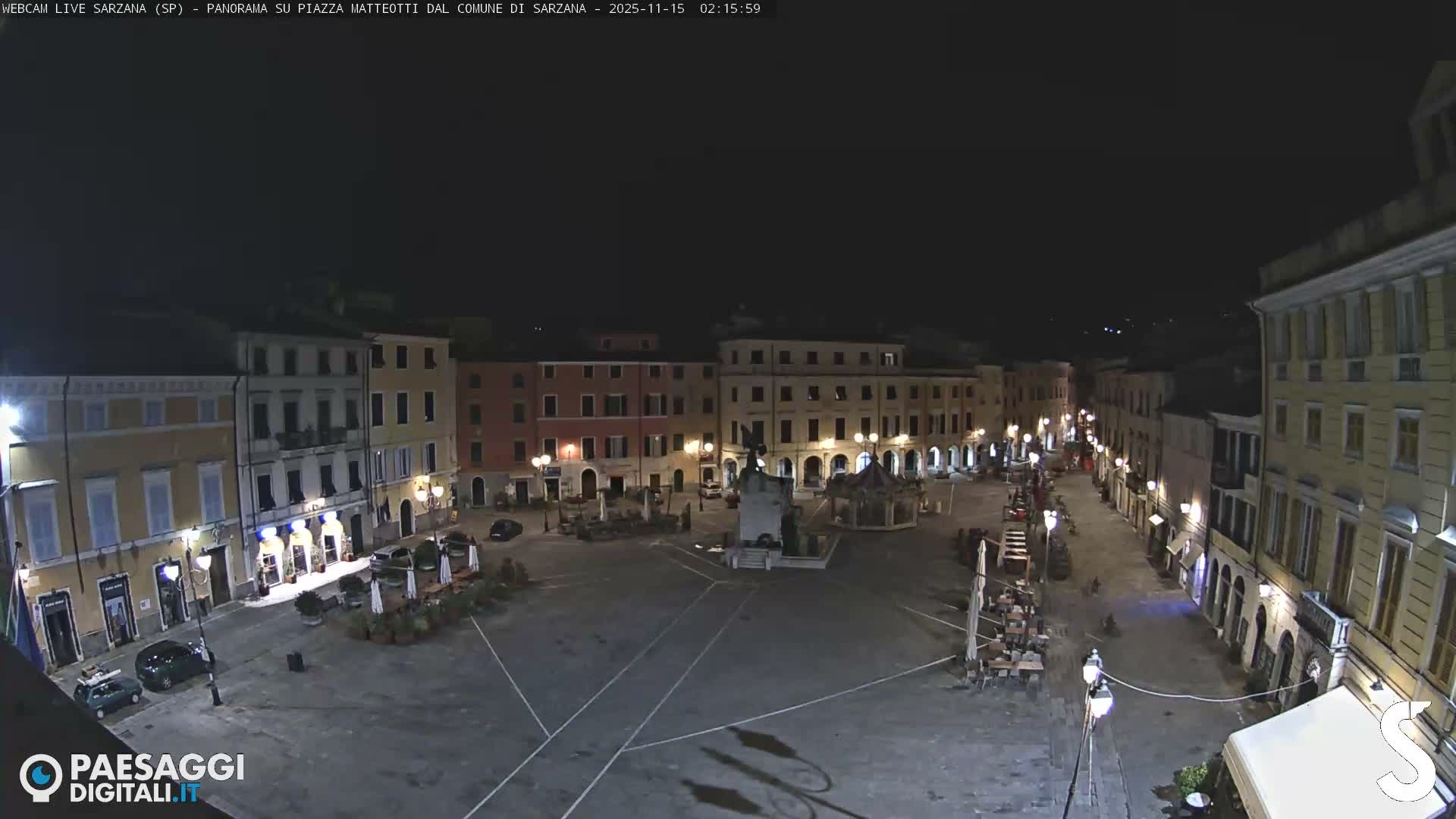 A wide view shows a quiet, well-lit town square at night, surrounded by colorful multi-story buildings, featuring a central monument, a gazebo-like structure, and outdoor seating areas under a clear sky.