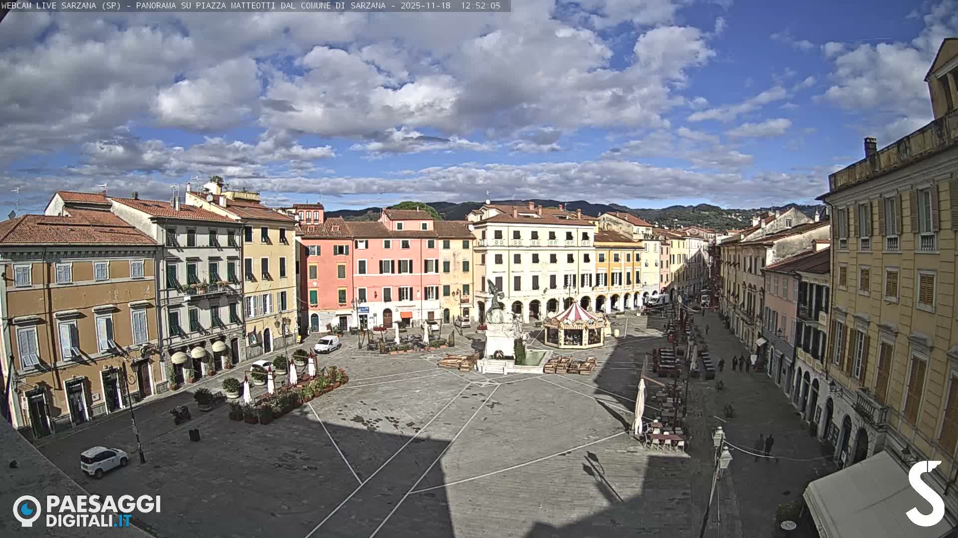 A wide view shows a quiet, well-lit town square at night, surrounded by colorful multi-story buildings, featuring a central monument, a gazebo-like structure, and outdoor seating areas under a clear sky.