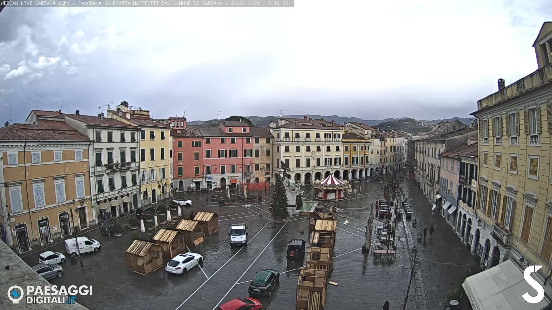 An overcast and wet town square is bustling with parked cars, pedestrians, wooden market stalls, a Christmas tree, and a carousel, all surrounded by colorful traditional European buildings under a grey sky with hills in the background.