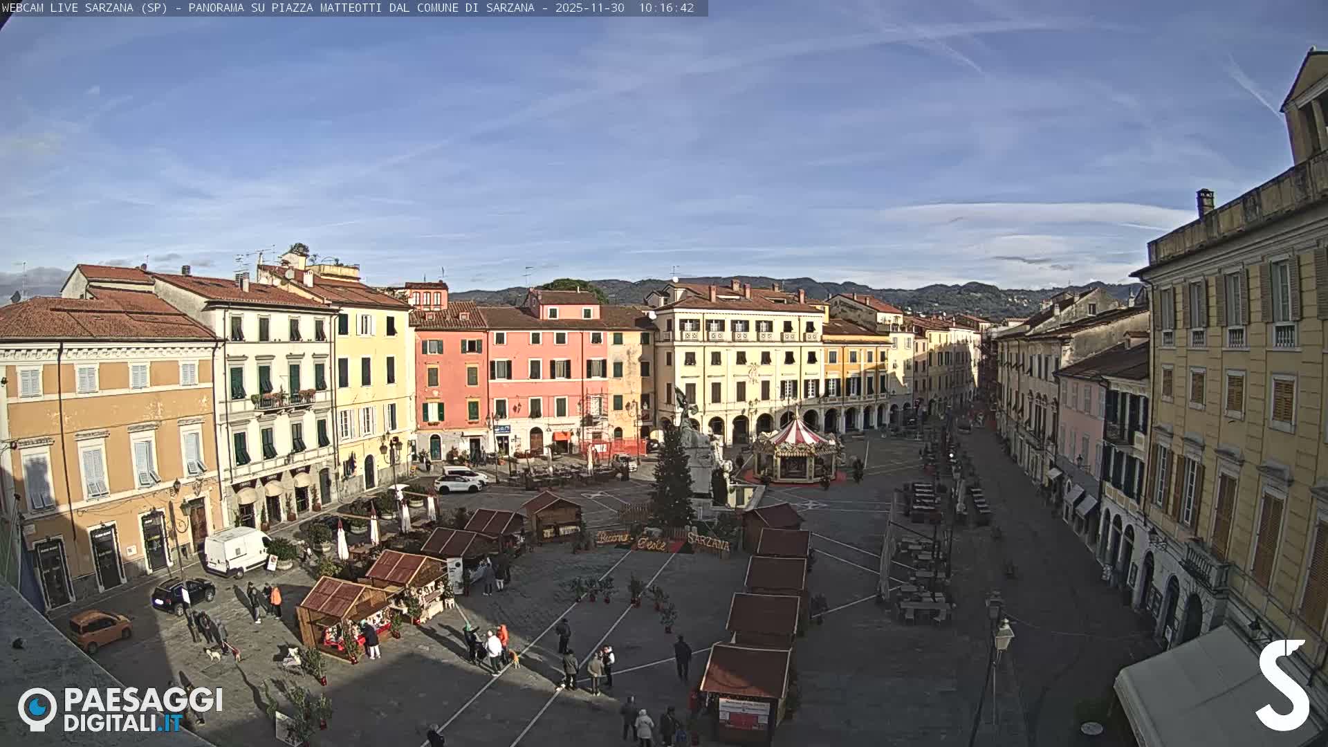 A vibrant European town square is seen on a clear, sunny day, featuring colorful buildings, a central Christmas tree, a carousel, and numerous market stalls with people strolling around.