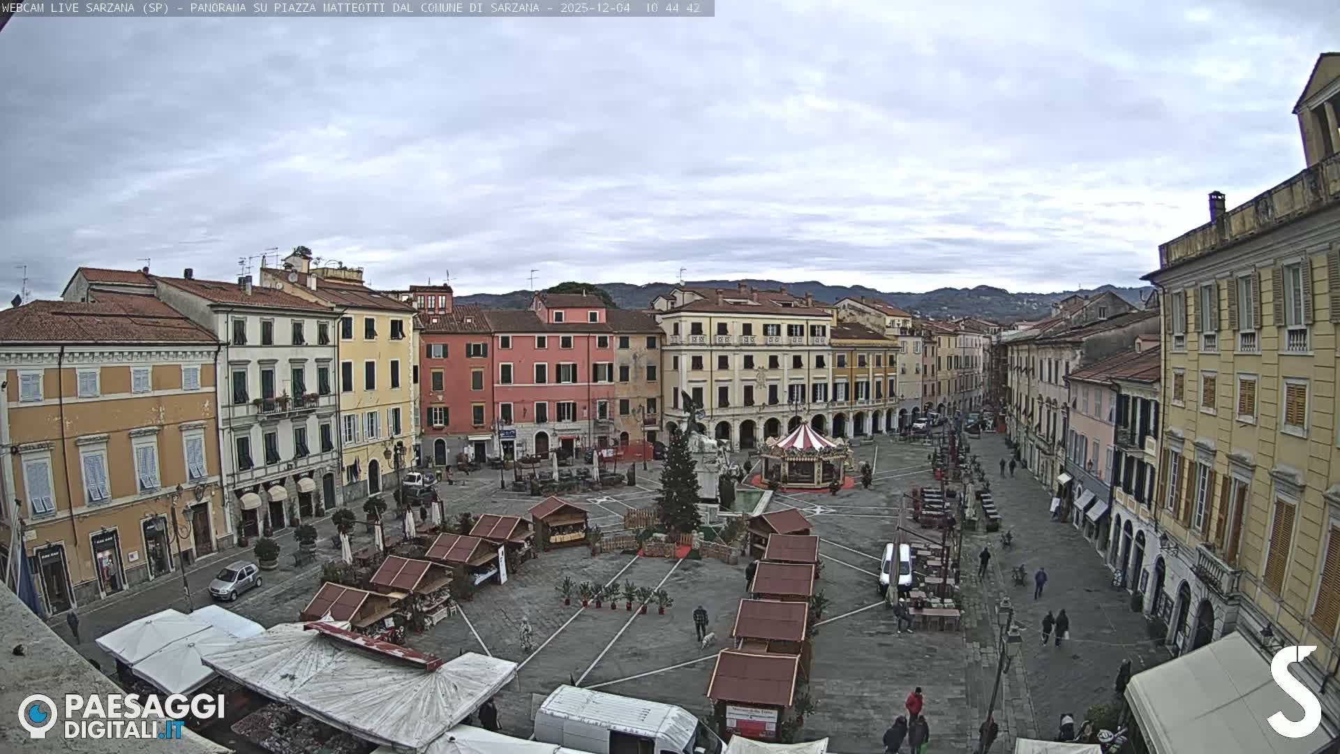 A wide view of a European town square on an overcast day shows colorful historic buildings, a decorated Christmas tree, a carousel, and numerous market stalls, with distant hills visible under a cloudy sky.