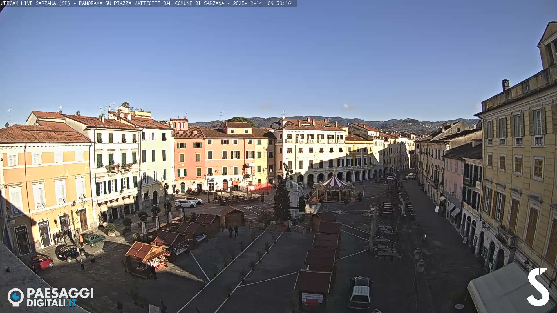 A wide view of a European town square on an overcast day shows colorful historic buildings, a decorated Christmas tree, a carousel, and numerous market stalls, with distant hills visible under a cloudy sky.