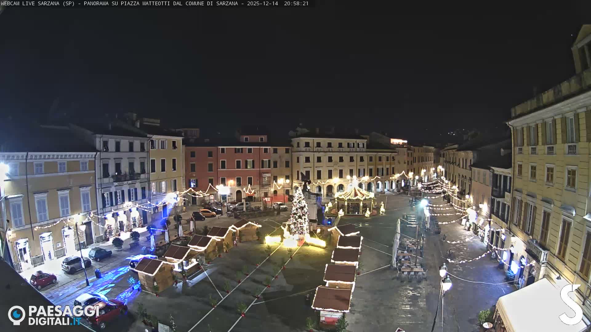 A wide view of a European town square on an overcast day shows colorful historic buildings, a decorated Christmas tree, a carousel, and numerous market stalls, with distant hills visible under a cloudy sky.