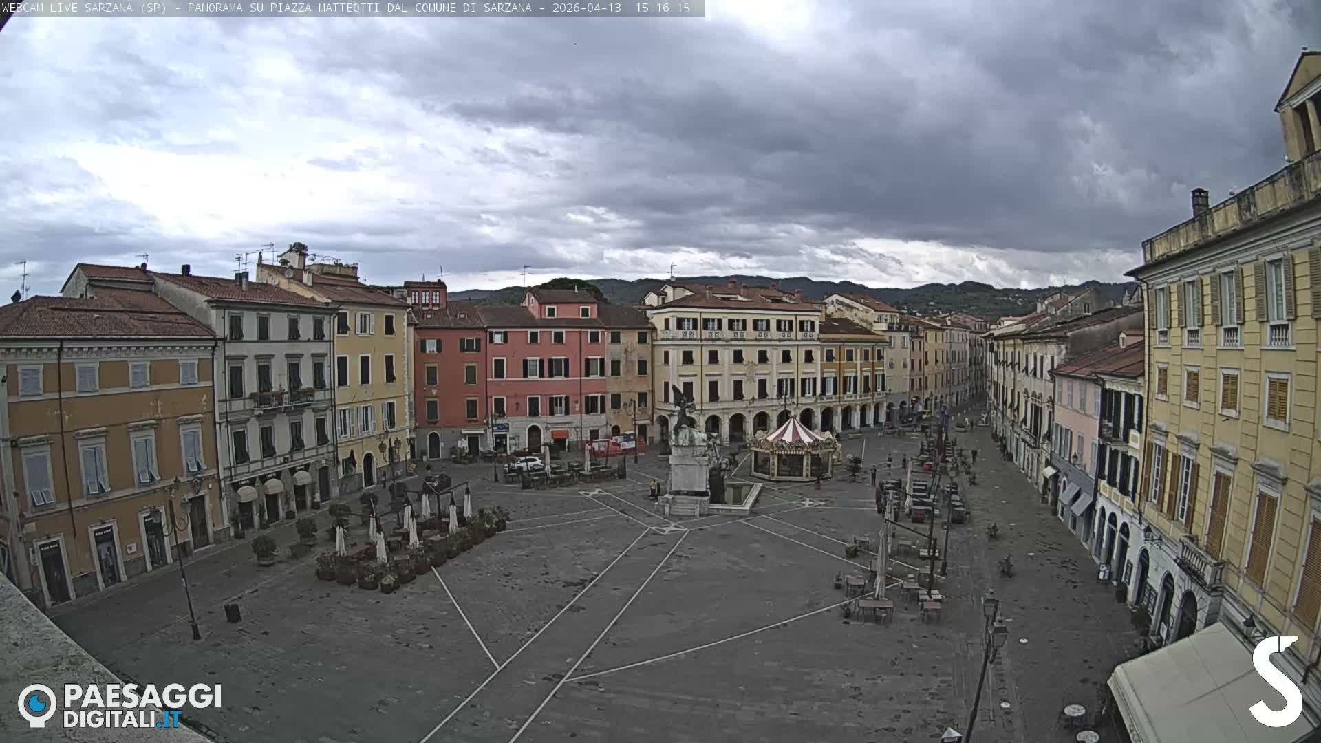 A wide view of a European town square on an overcast day shows colorful historic buildings, a decorated Christmas tree, a carousel, and numerous market stalls, with distant hills visible under a cloudy sky.