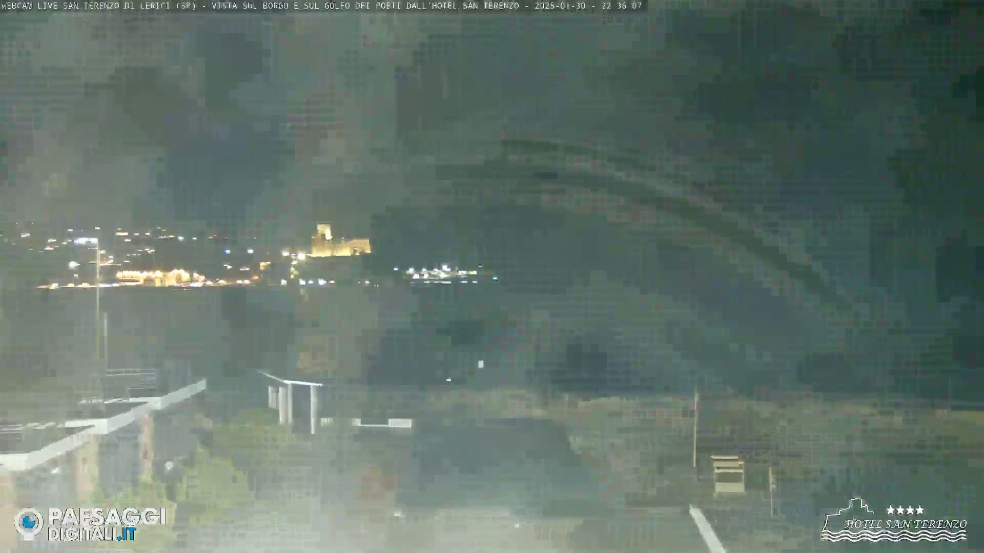 A nighttime long shot shows a blurry, hazy view of a town's lights across a body of water, under a dark, overcast sky.