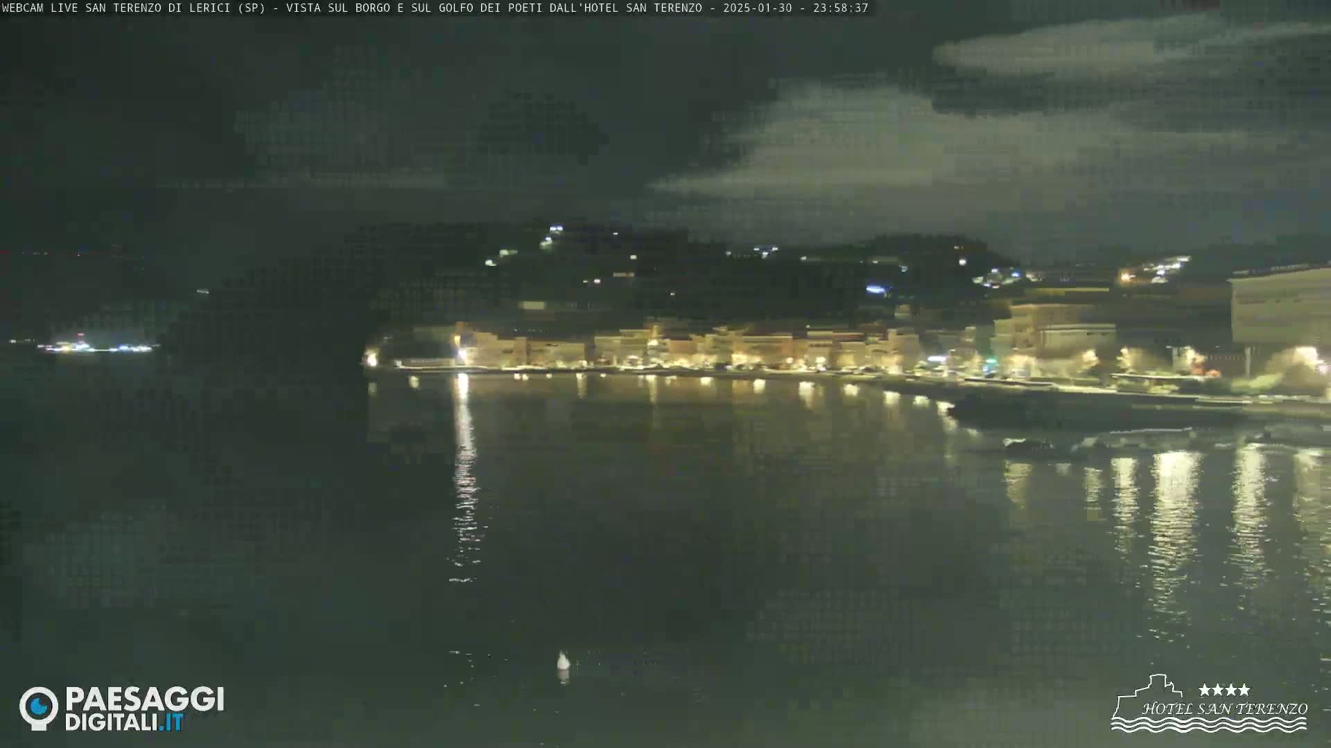A nighttime view of a calm body of water reflecting the lights of a town under a partly cloudy sky.