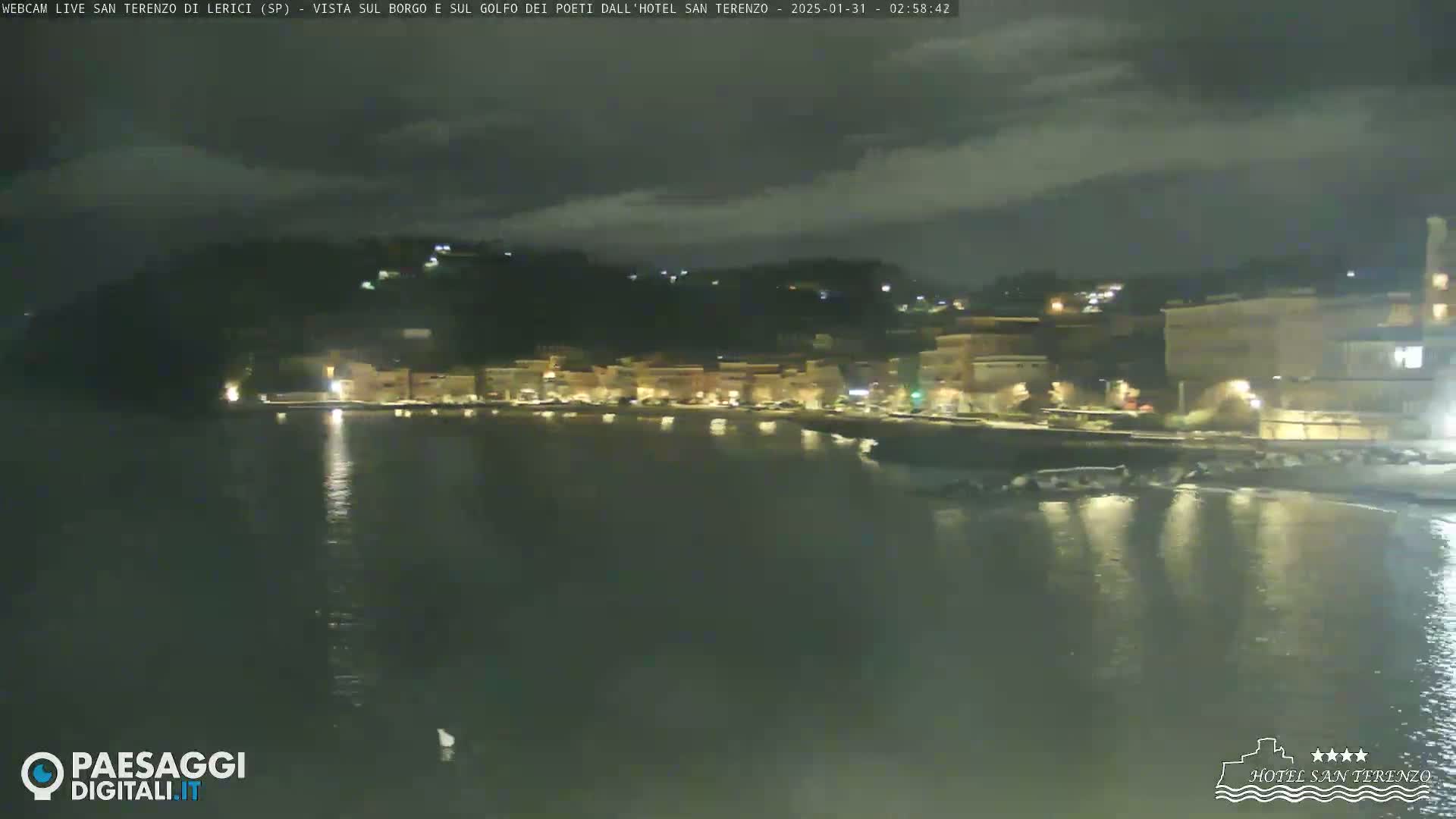 A nighttime view of a town's waterfront, illuminated by streetlights, under a partly cloudy sky.