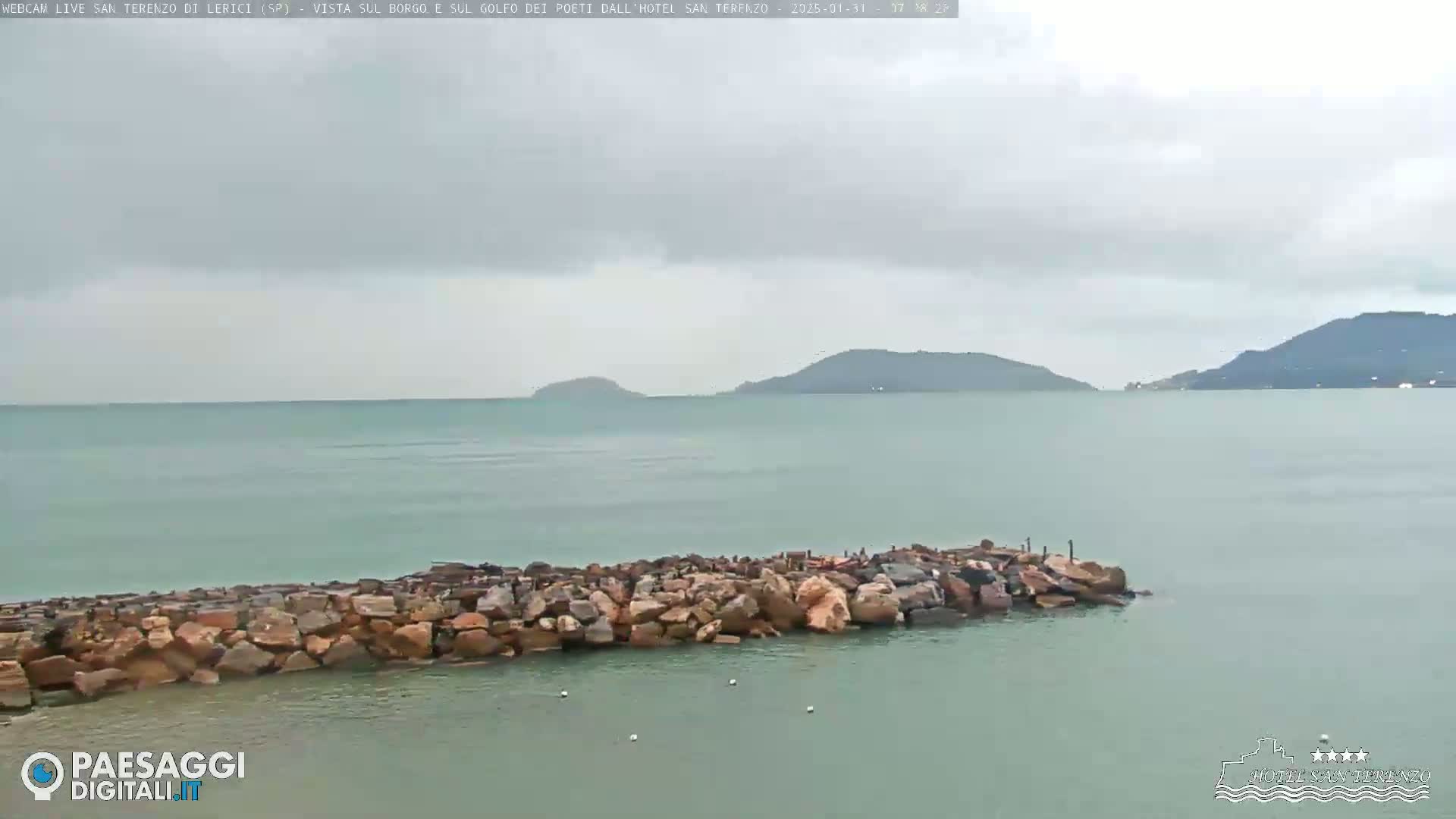 A rocky breakwater extends into a calm, green-grey sea under an overcast sky, with distant hills visible on the horizon.
