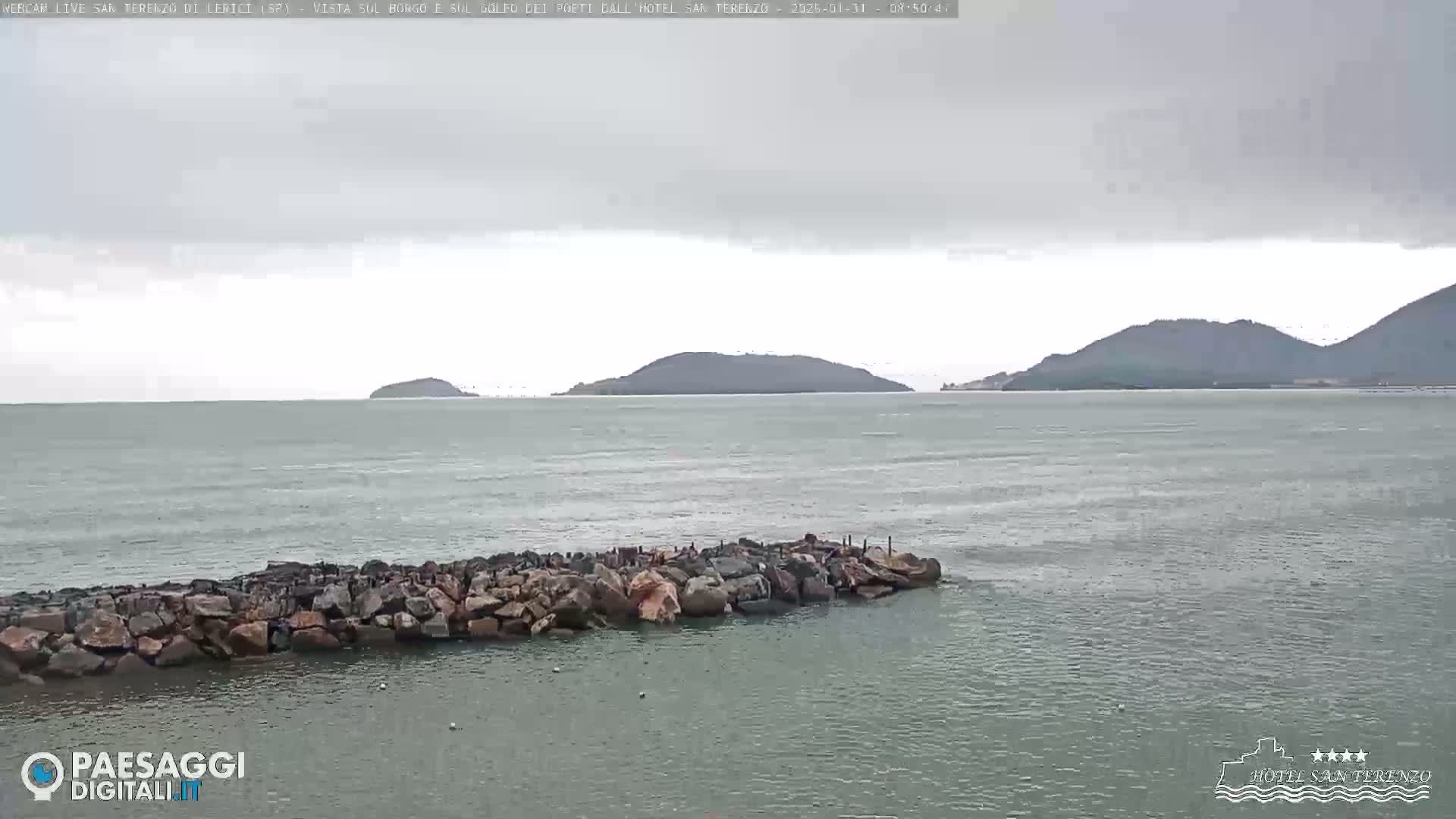 A rocky breakwater extends into a calm, gray sea under an overcast sky, with several distant hills visible on the horizon.