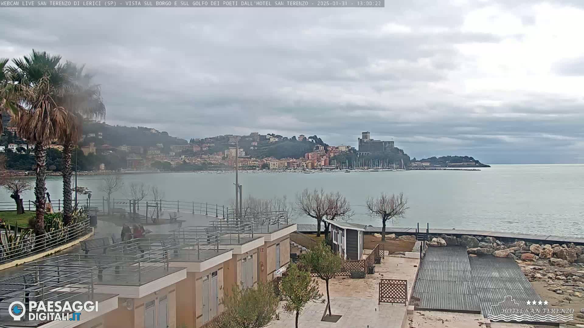 A coastal town with a castle is visible across a calm body of water under an overcast sky, viewed from a waterfront area with modern buildings and landscaping.