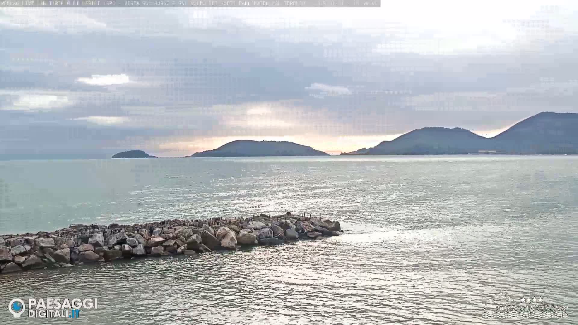 A rocky breakwater extends into a calm sea under a mostly cloudy sky, with distant hills visible on the horizon.
