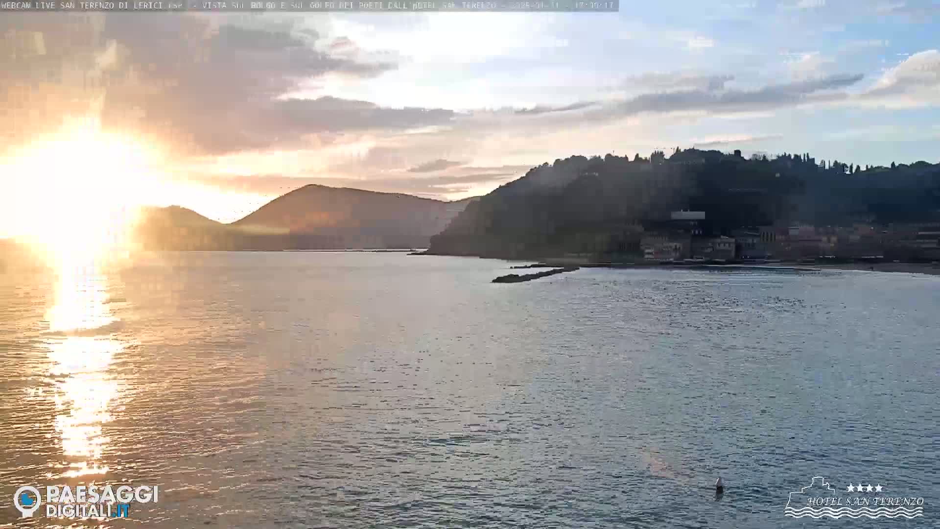A calm body of water reflects the setting sun, with hills and a town visible in the background under a partly cloudy sky.
